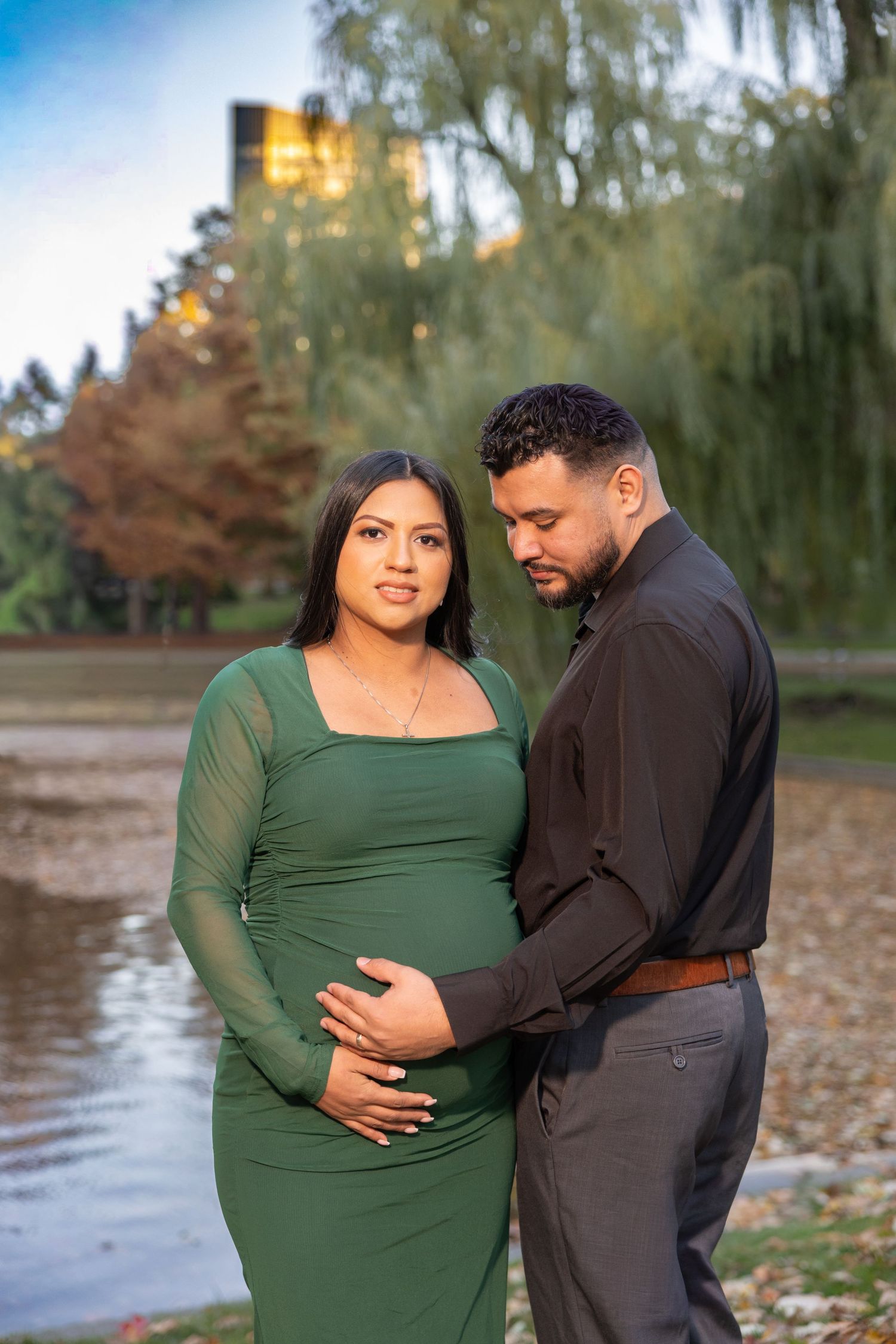 A couple in coordinating green and black outfits poses together outdoors near a lake during autumn.