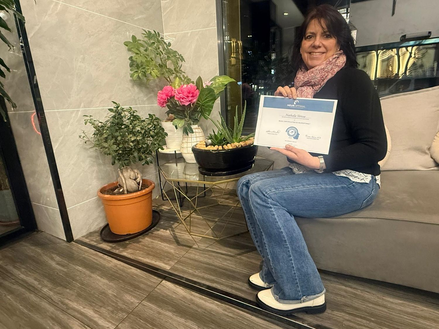 Someone sits on a porch with a certificate while surrounded by potted plants including pink flowers.