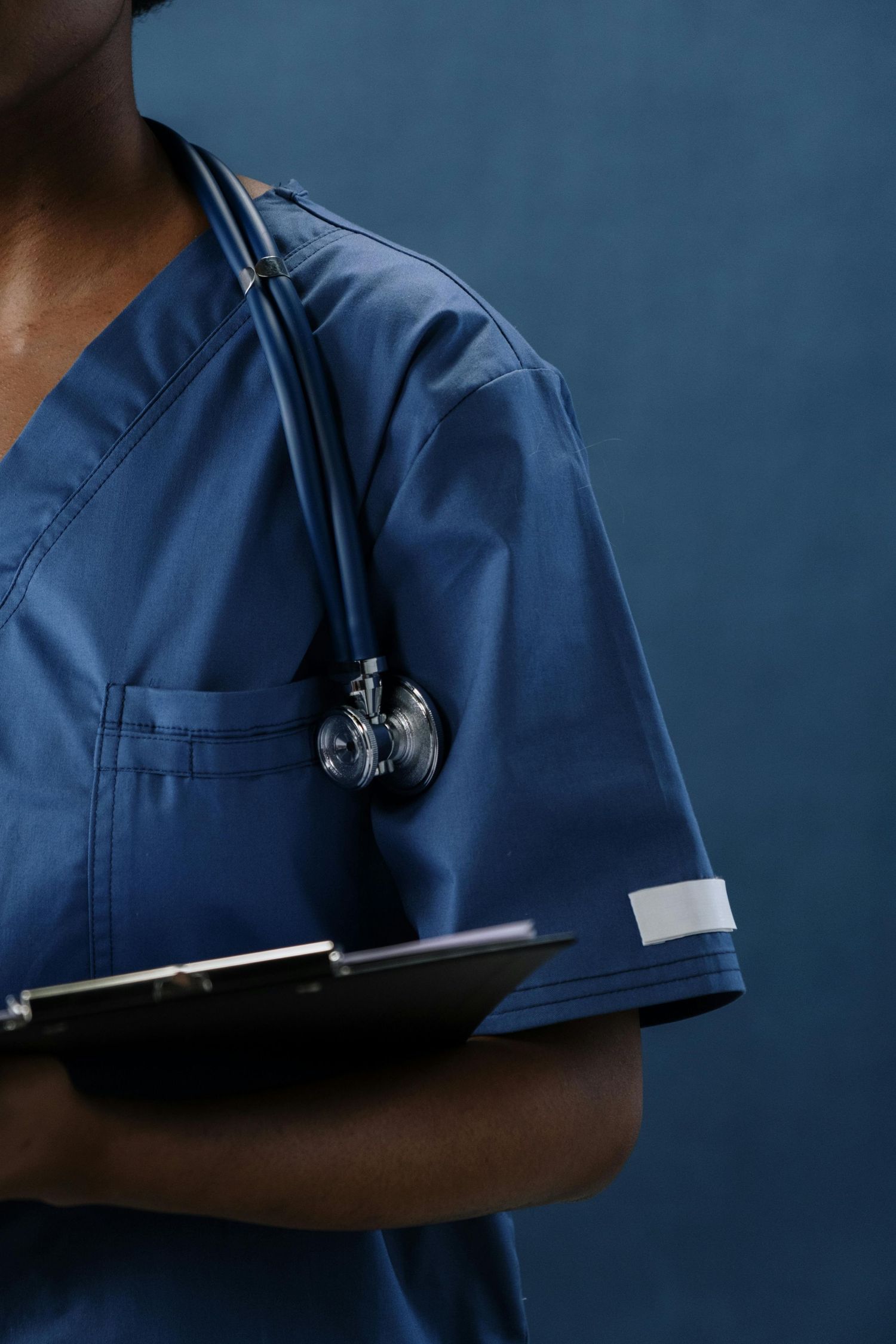 Healthcare professional in blue scrubs holds clipboard while wearing stethoscope against dark background.
