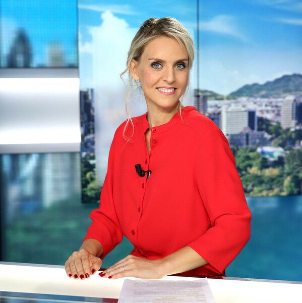 A broadcast presenter in a vibrant red blazer sits at a news desk with a city skyline visible in the background.