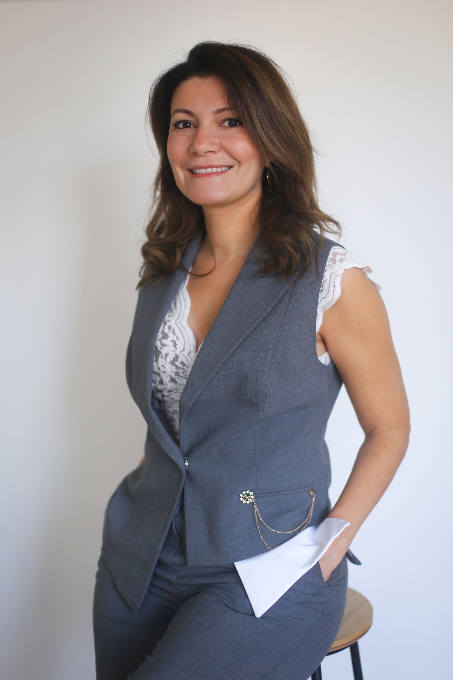 Professional photo of someone wearing a gray business suit vest with white lace blouse in a minimalist setting.
