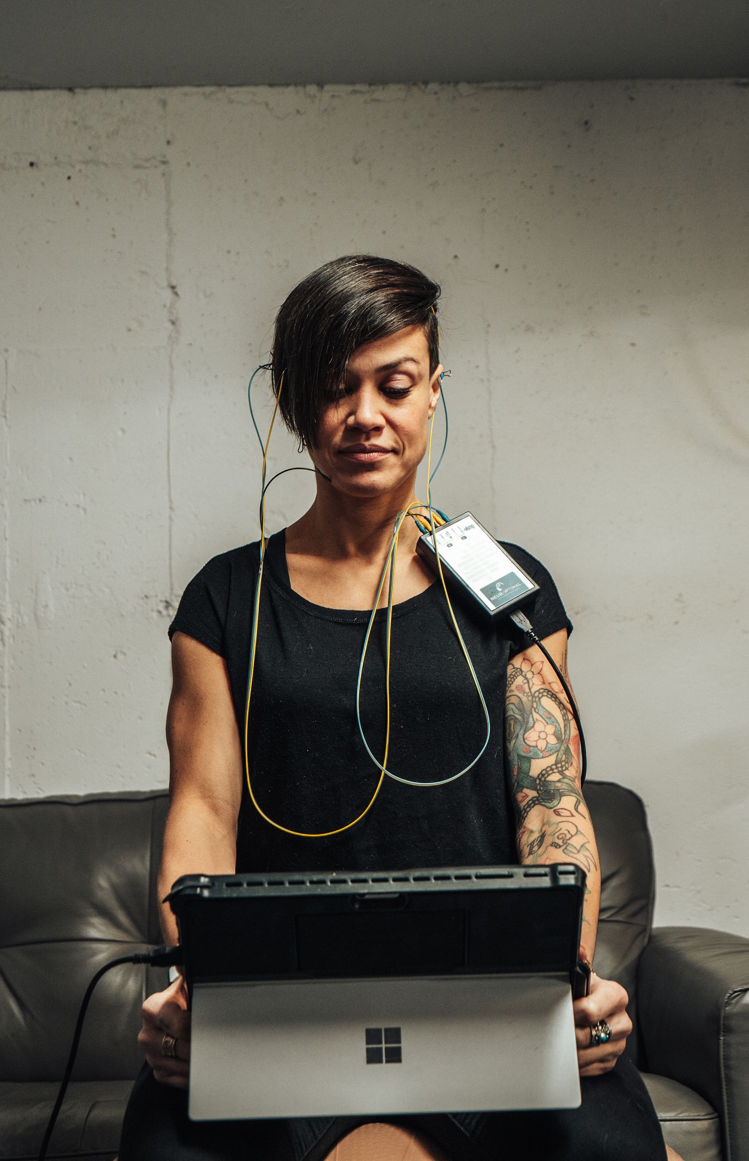 Person with tattoos wearing black tank top works on Microsoft Surface laptop while wearing yellow headphones.