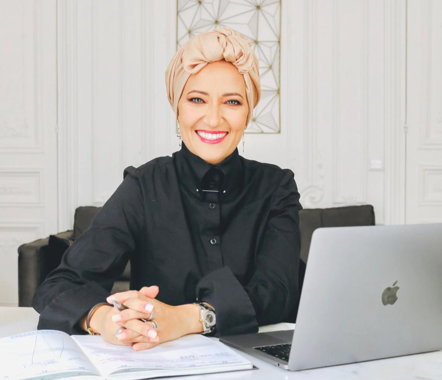 A professional in a black blazer sits at a desk with a MacBook laptop and paperwork in a bright modern office space.