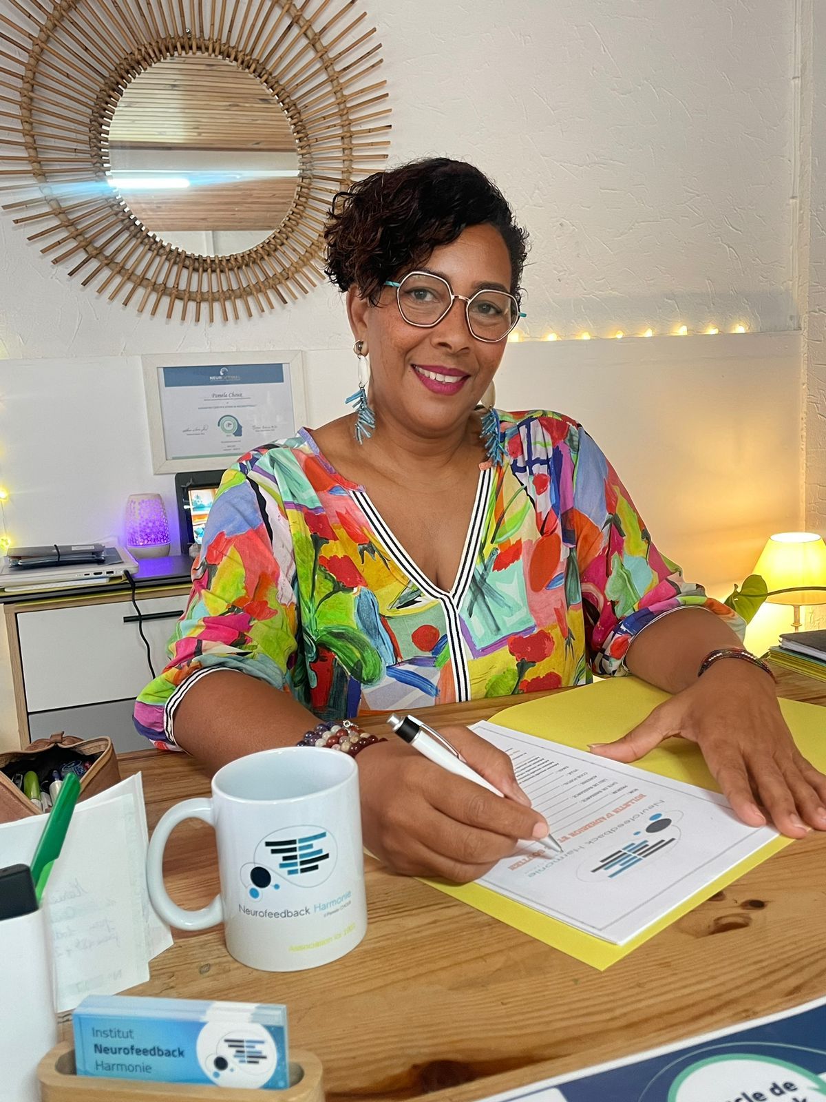 Professional at wooden desk wearing colorful floral print dress and glasses writes on document with coffee mug nearby.