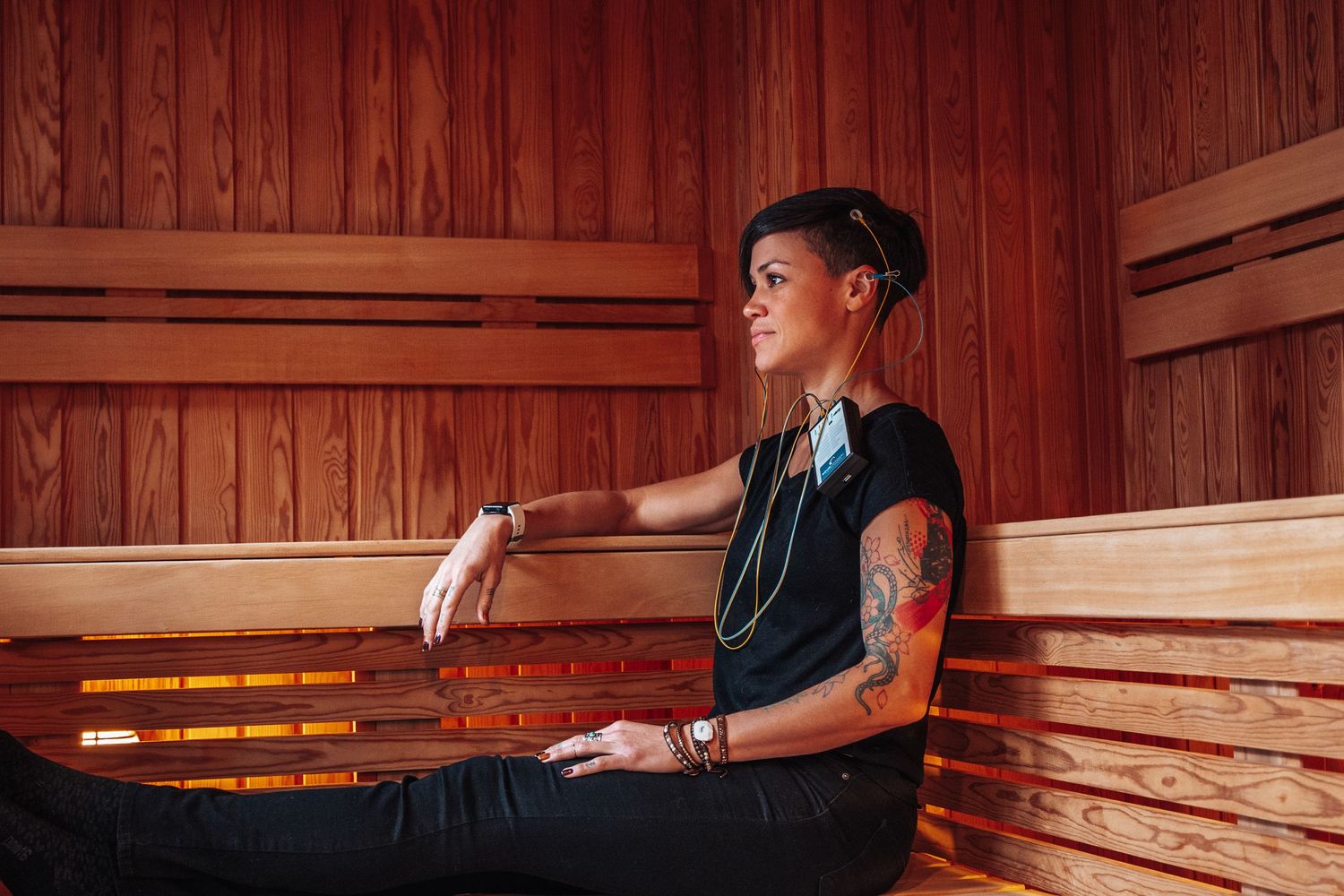 Someone relaxes in a wooden sauna while wearing a black tank top and listening to music with earbuds.