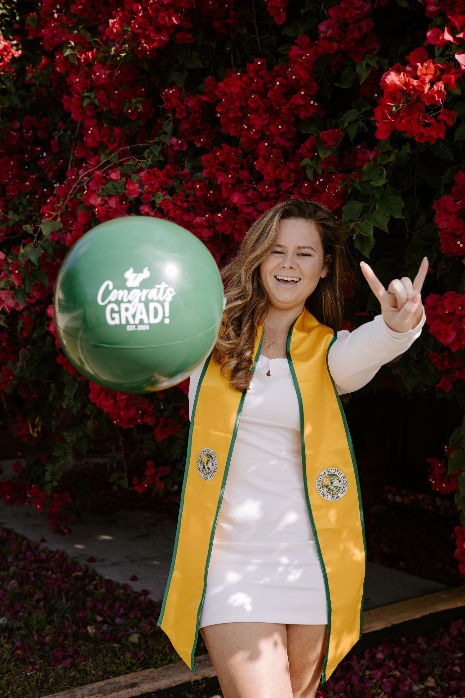 Graduate in yellow stole poses with congratulatory balloons against red flowering bush.