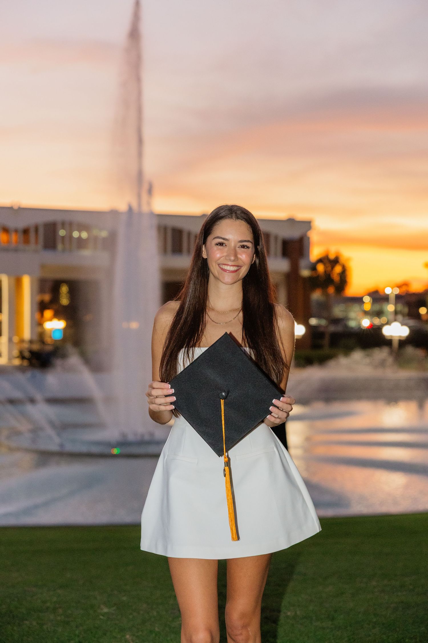 Graduate in cap and gown poses outdoors during golden sunset with university buildings in background.
