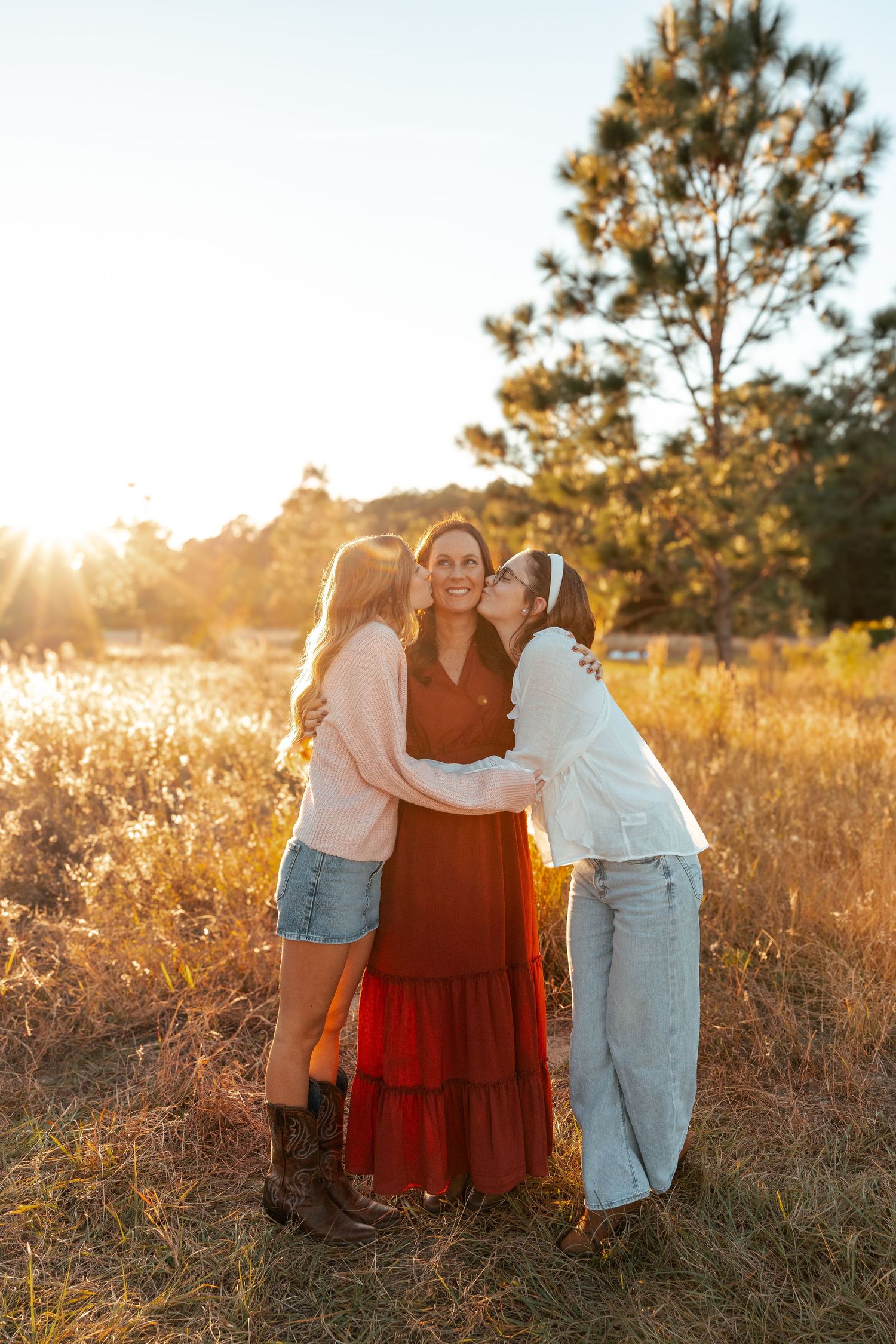 Three people share a tender embrace in a sunlit field with tall grass and pine trees at golden hour.