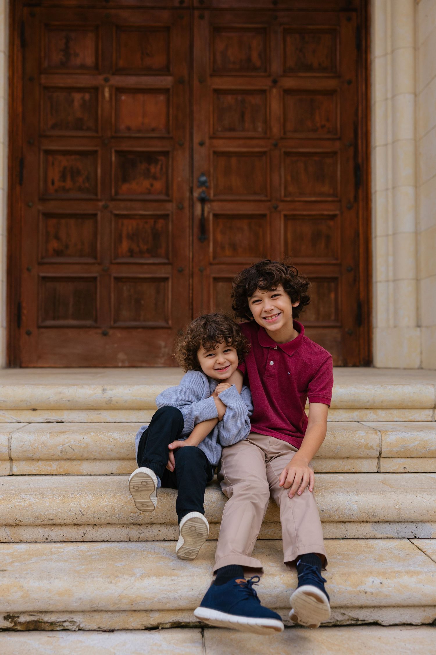 Two children sit together on stone steps in front of large wooden doors, sharing a playful moment.