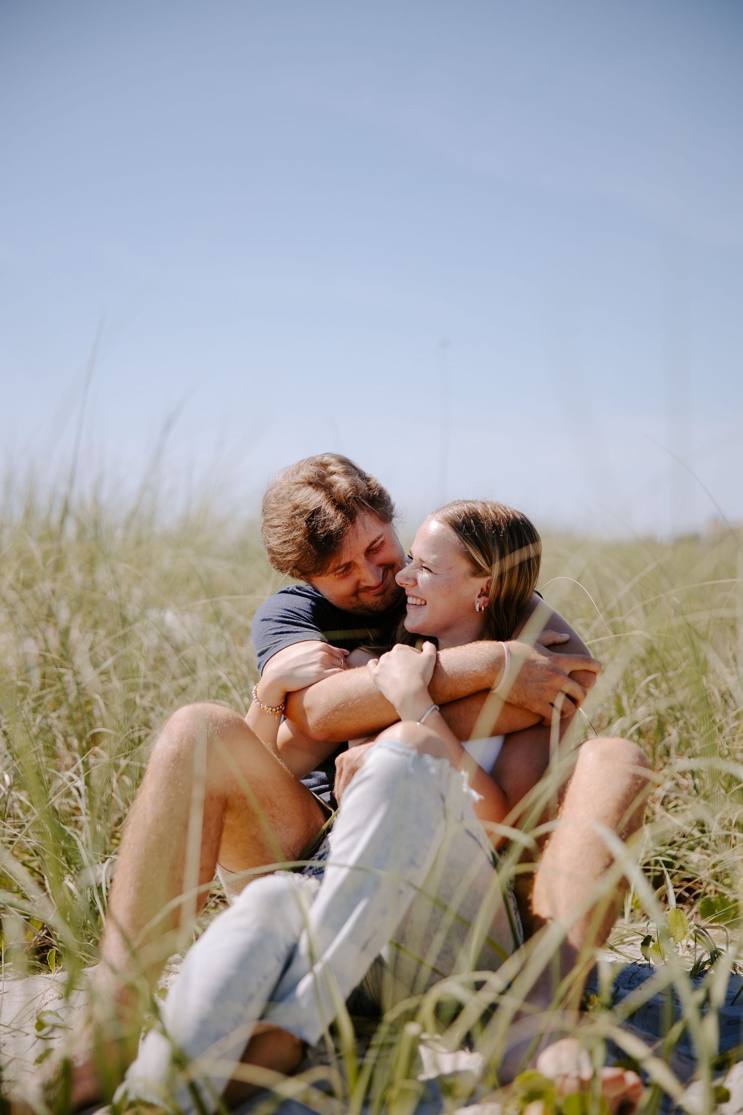 A romantic couple sits together in tall grass on a sunny day, sharing intimate moments in a natural setting.