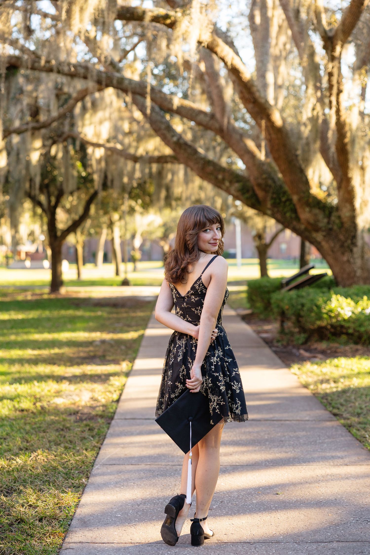 A floral dress-clad figure poses on a sunlit path beneath Spanish moss-draped oak trees in a park setting.