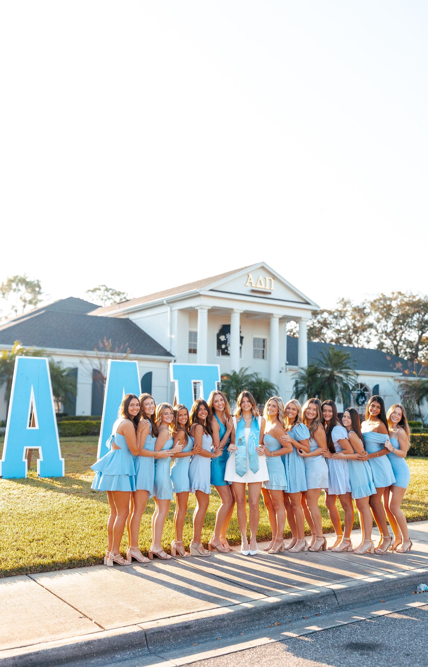 A group in matching light blue dresses poses together in front of large blue letters and a white columned building.