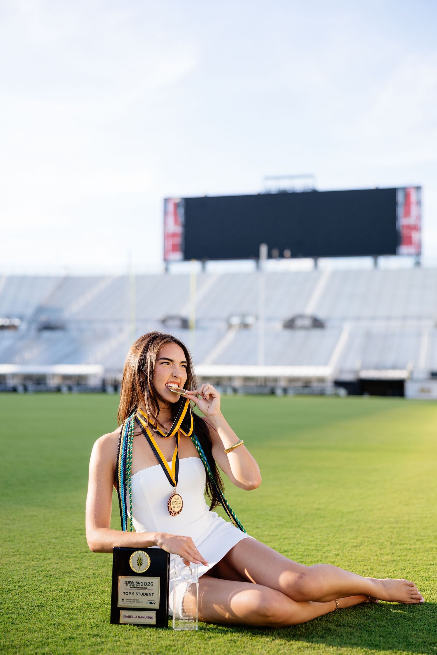 Graduate in white dress sits on stadium field with awards, wearing green and gold honor cords, biting a medal.
