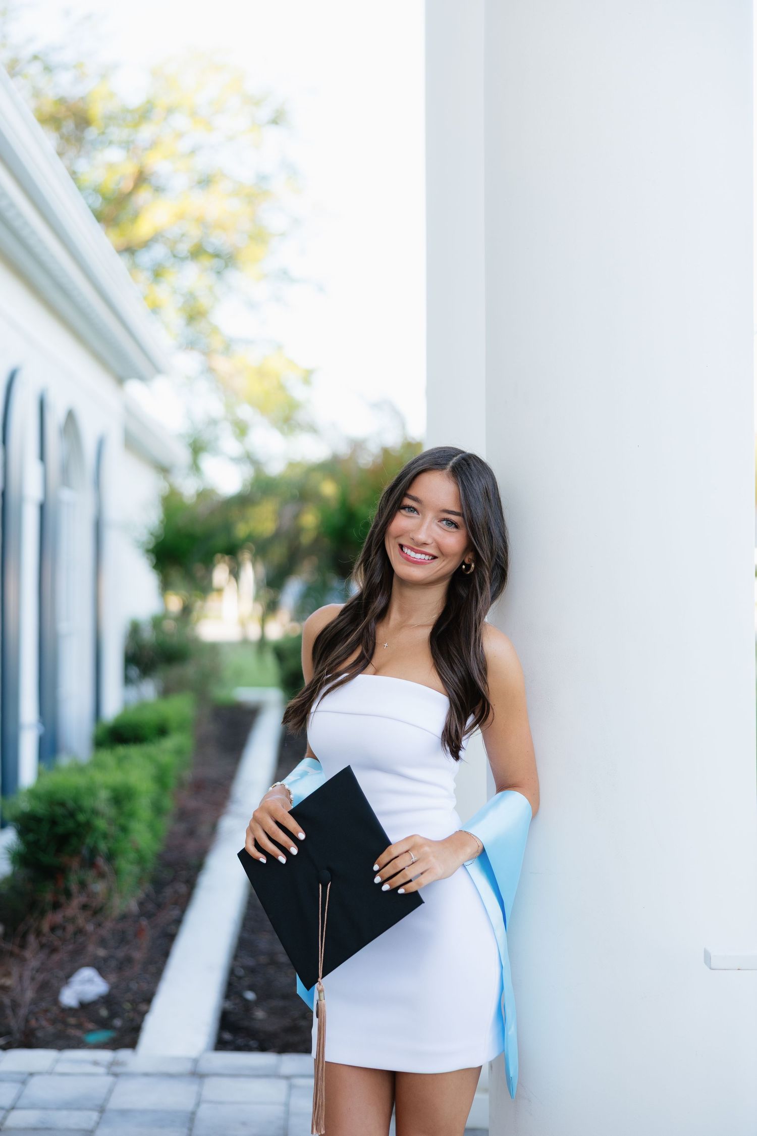 Smiling young woman in white strapless dress holding graduation diploma and cap outdoors near white building.