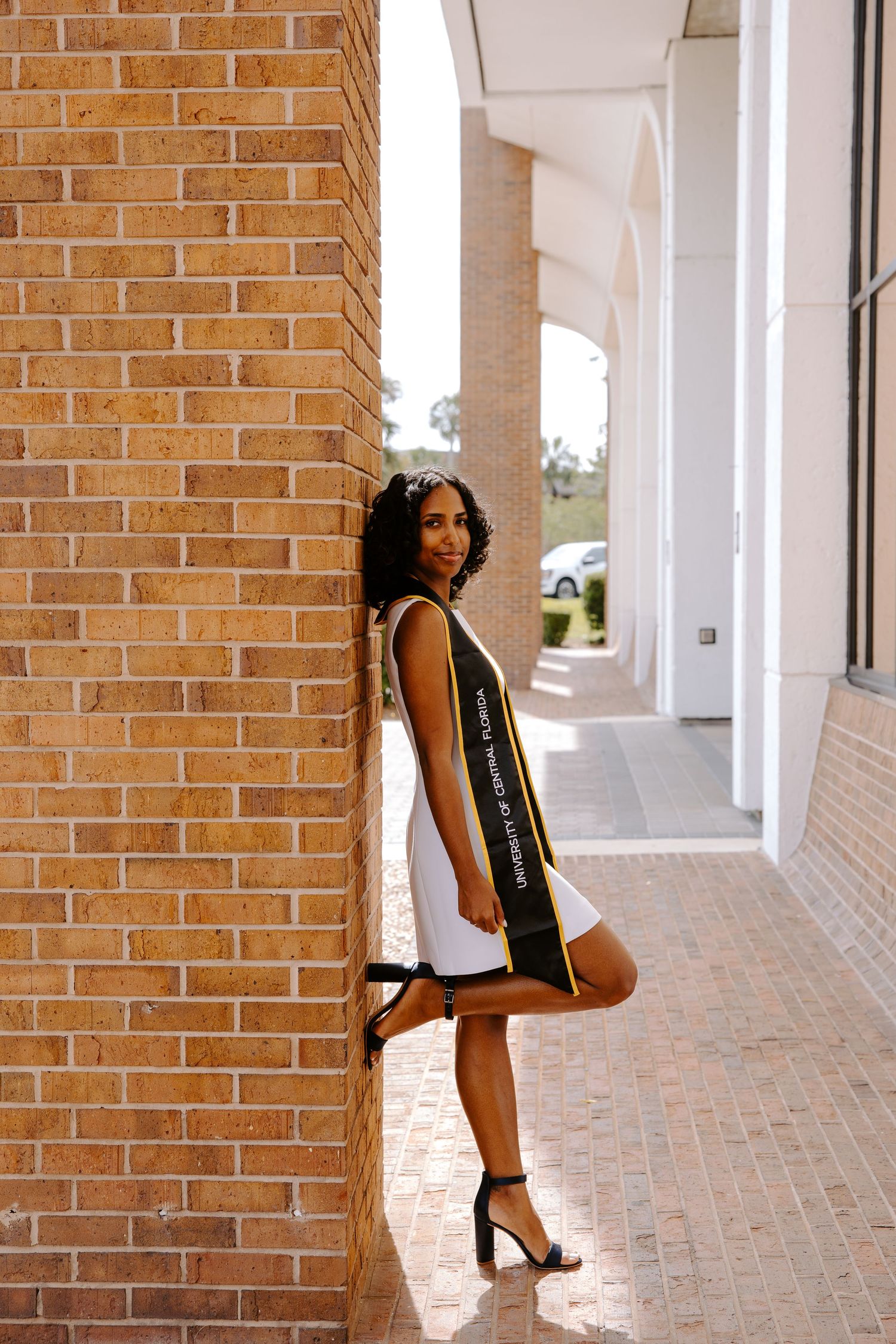 Graduate in cap and gown poses playfully against brick columns.