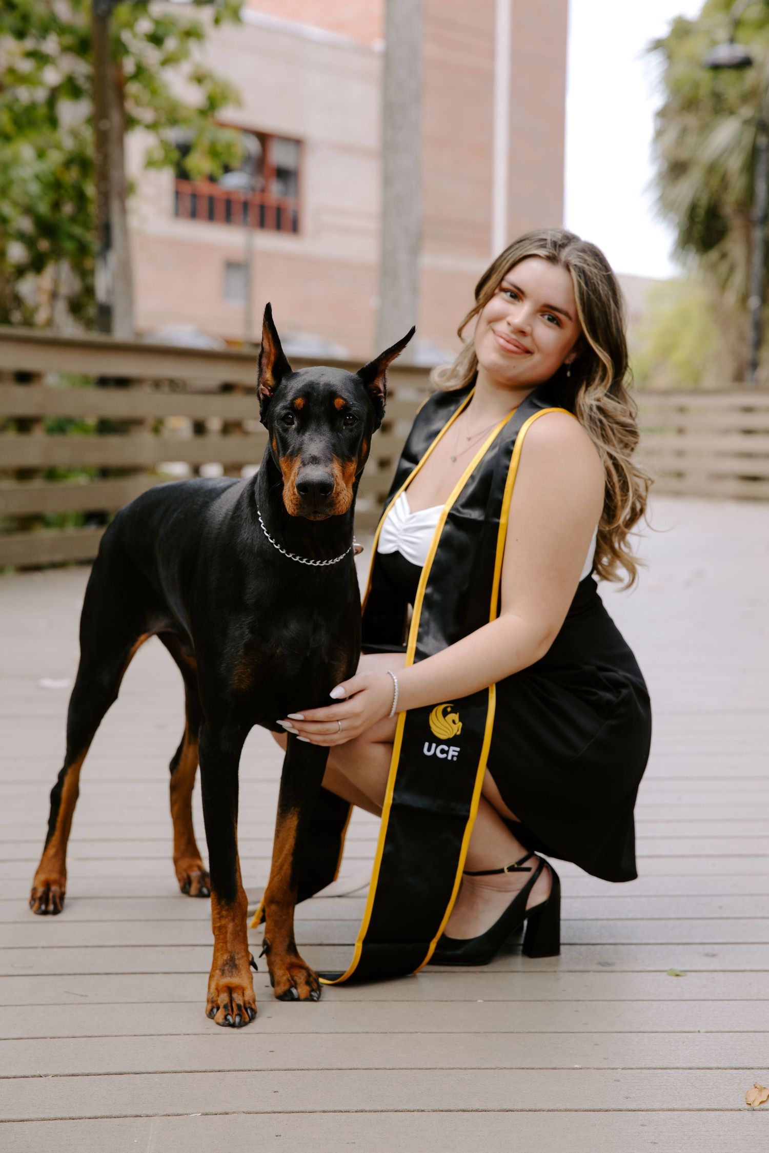 Graduate poses in black dress with graduation sash beside a Doberman Pinscher on wooden deck.