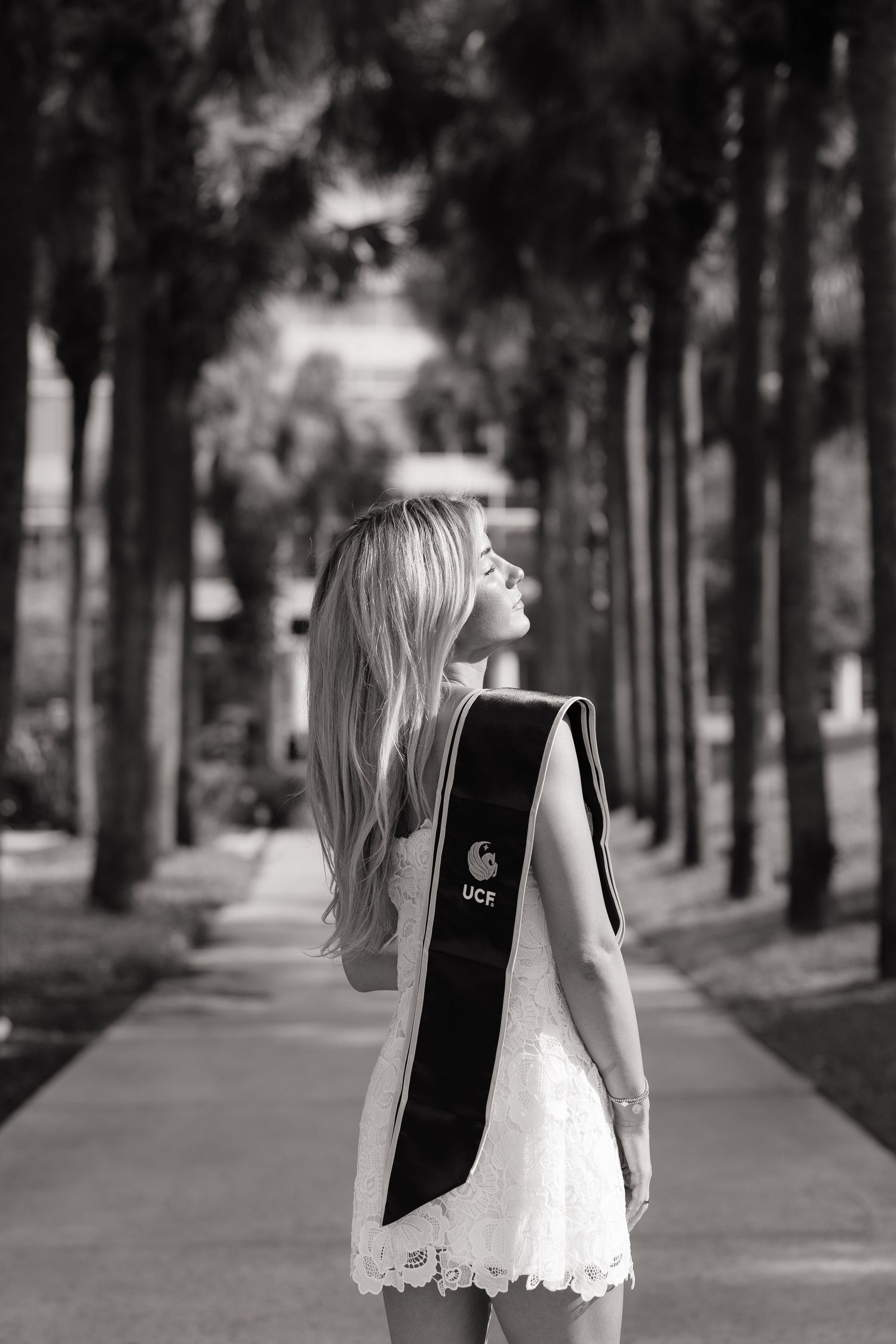 Black and white photo of a blonde woman in a white dress wearing a UCF graduation sash among palm trees.