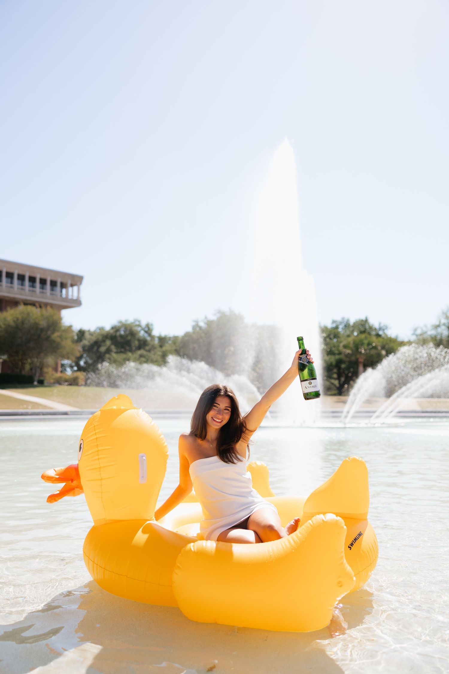 Woman sitting on giant inflatable yellow duck in fountain, holding beer bottle up celebrating on sunny day.
