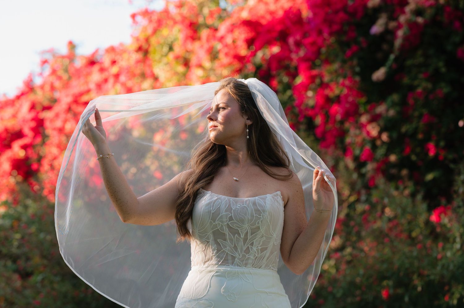 A bride in a white gown poses gracefully against a backdrop of vibrant red bougainvillea flowers in a garden setting.