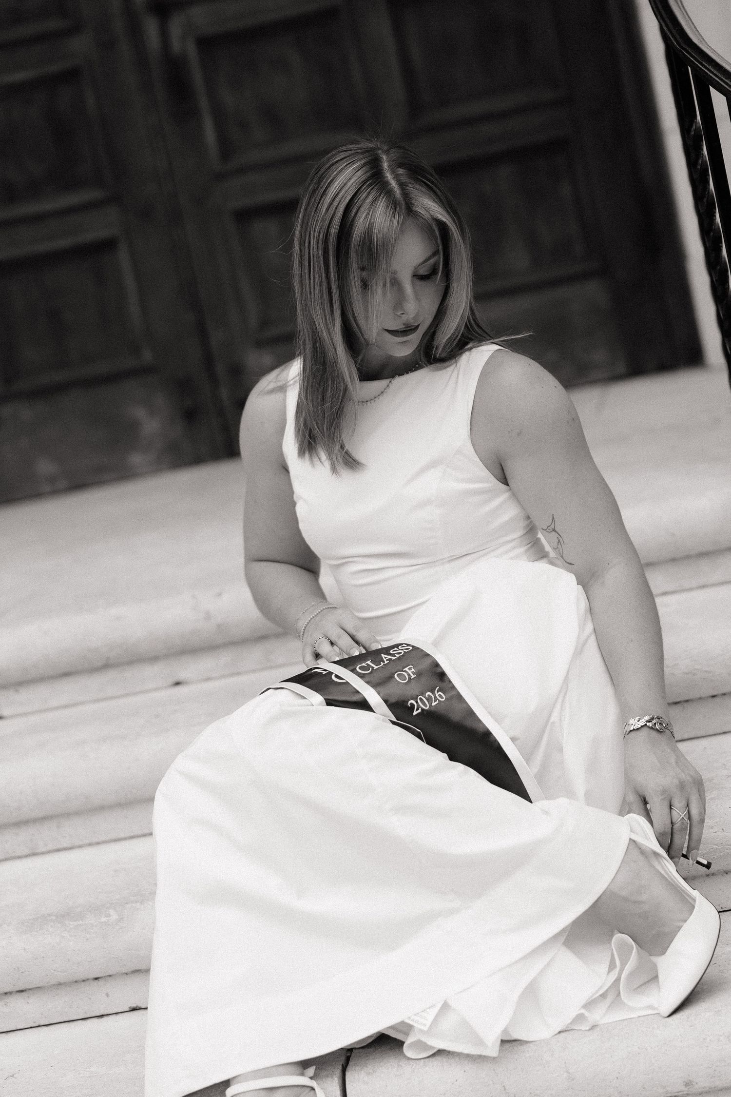 Black and white photo of a young woman in a white dress wearing a Class of 2026 sash, sitting on steps.