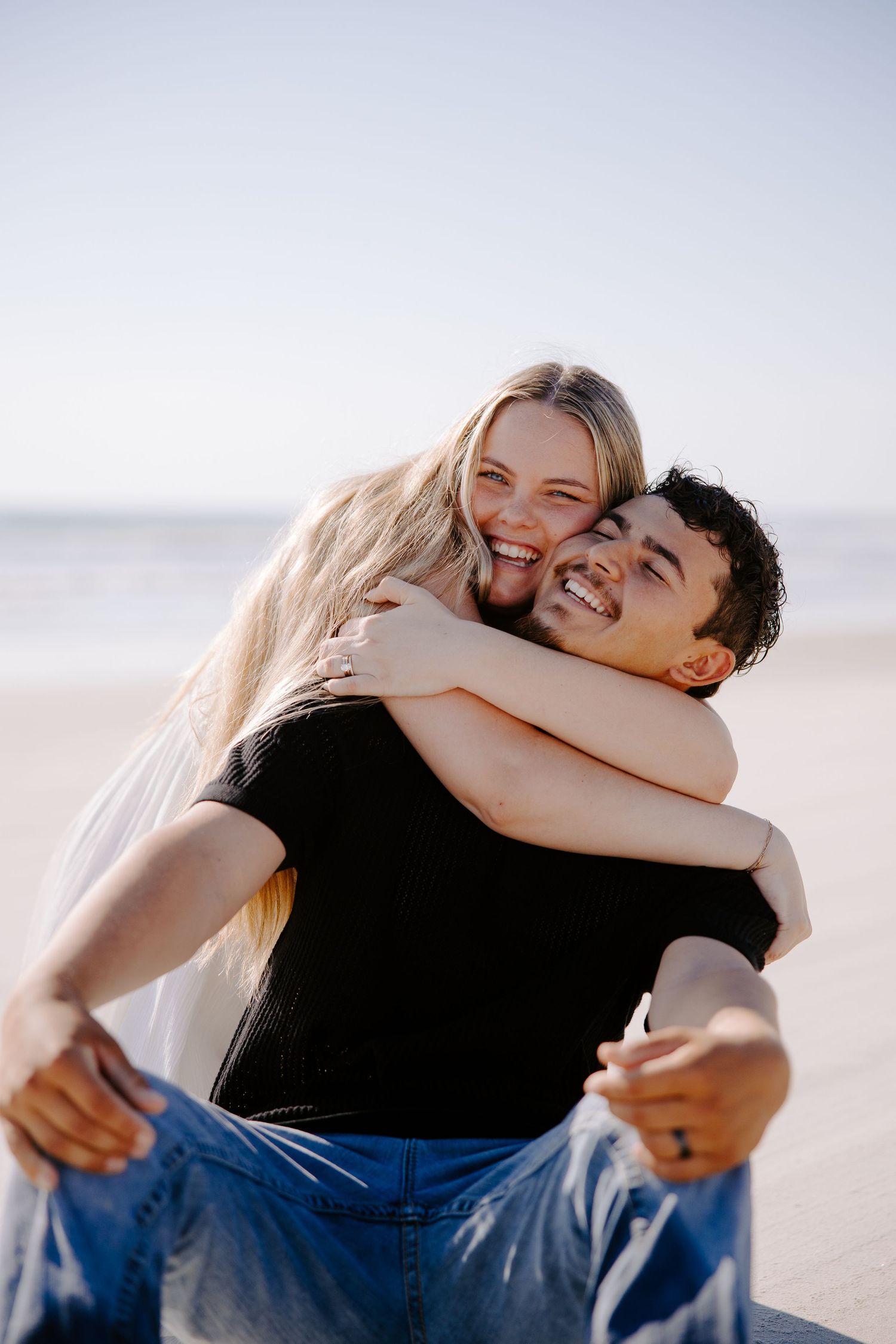 Happy couple laughing and embracing on a sunny beach with clear sky in the background.