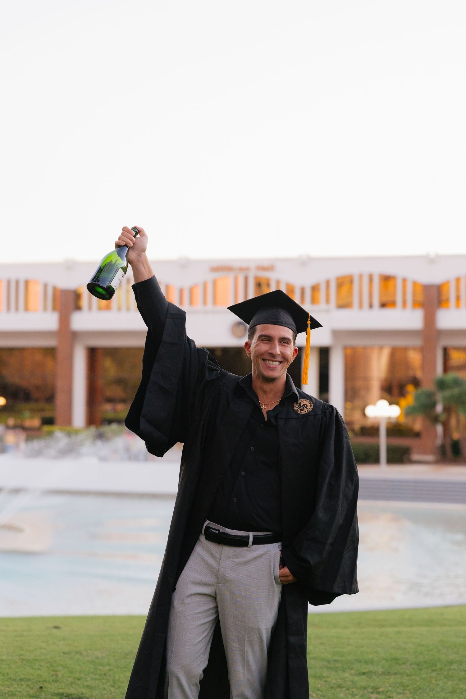 A graduate in cap and gown celebrates on a lawn with a champagne bottle during a sunny graduation photoshoot.