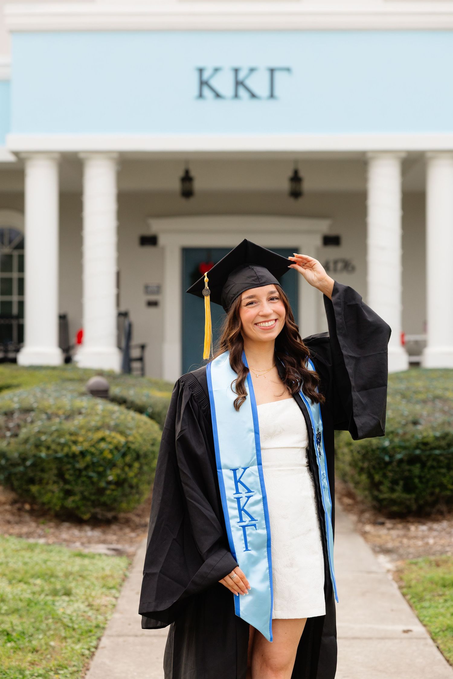 Graduate in cap and gown smiling in front of white pillared building during graduation photo session.