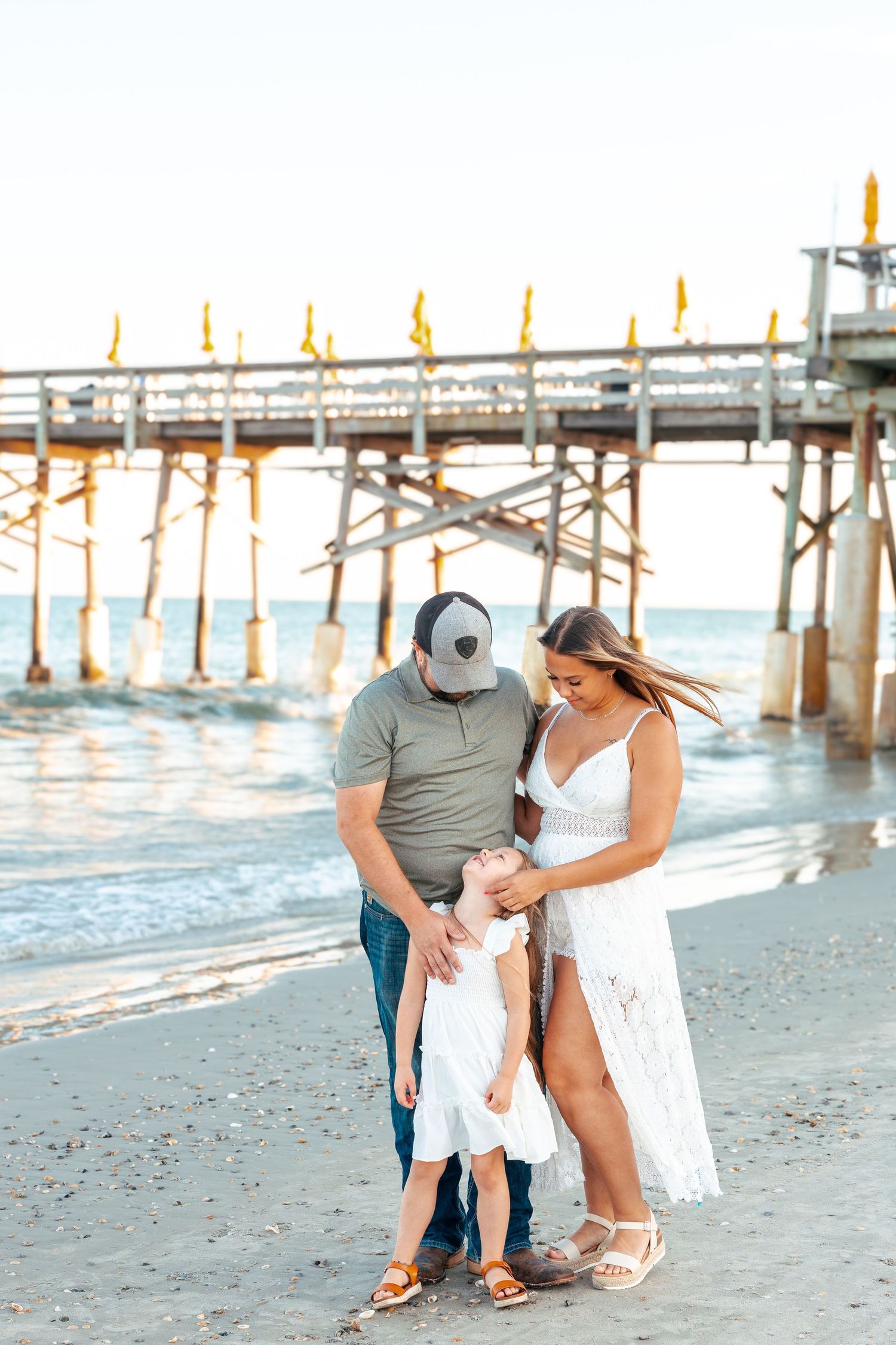 Family in white outfits sharing tender moments on a beach with wooden pier in background at sunset.