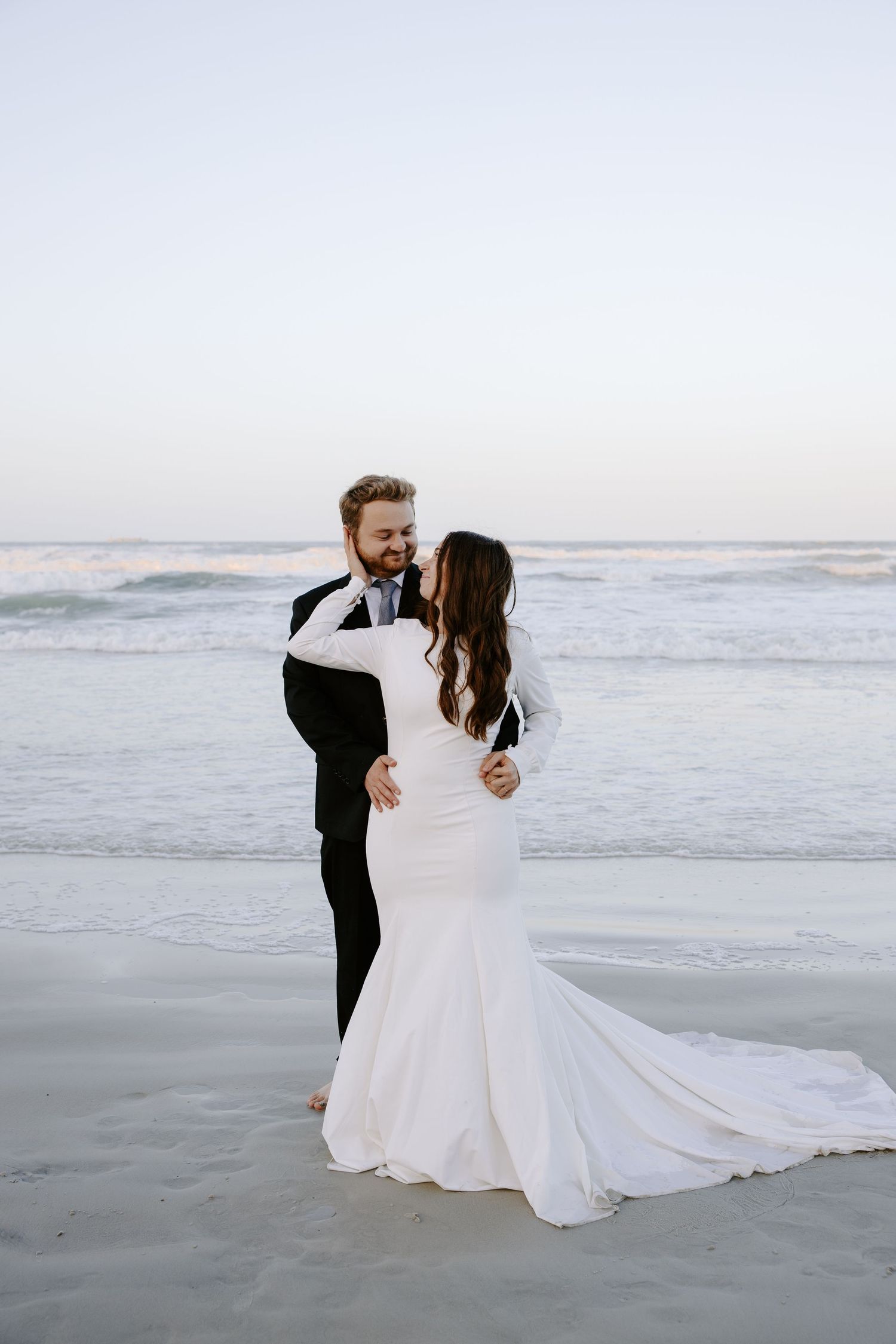 A romantic wedding couple dances and embraces on a misty beach at sunset in a flowing white gown and black suit.