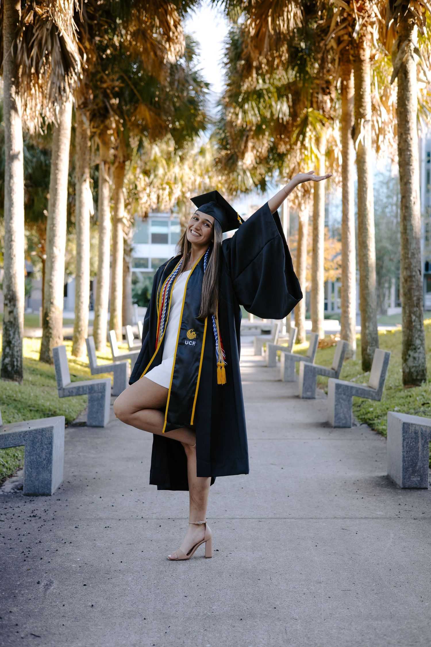 Graduate in cap and gown posing between palm trees on campus walkway.