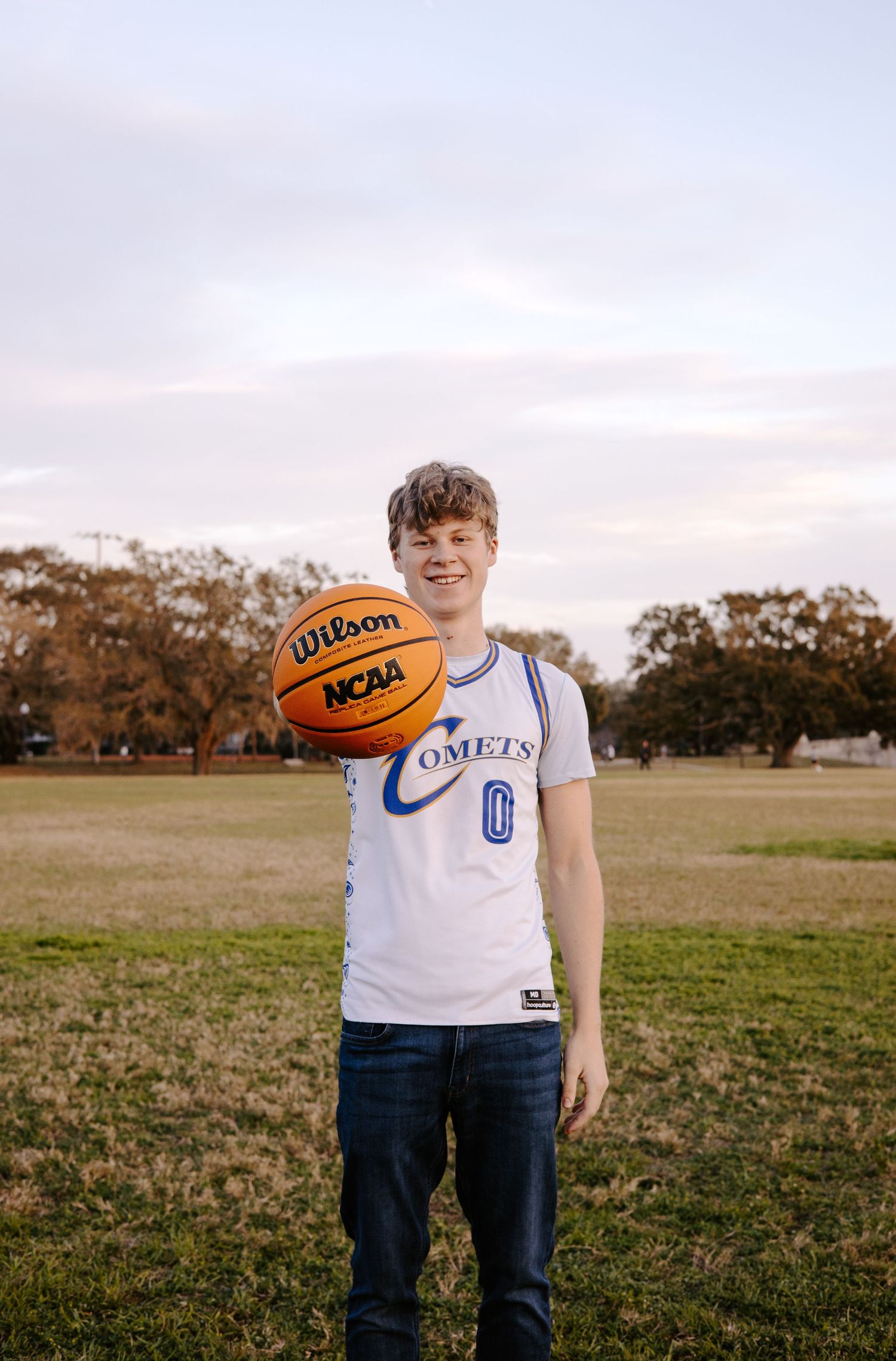 Graduate in UCF shirt holds basketball while posing in open field at sunset.