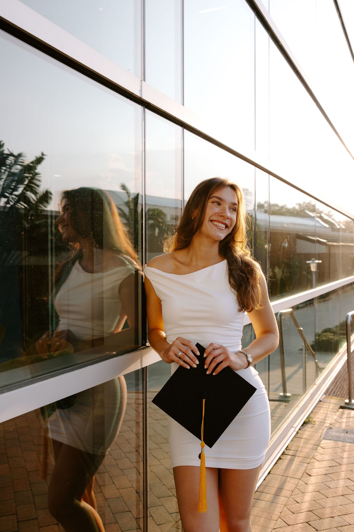 Graduate in white top with graduation stole poses against reflective glass windows at sunset.