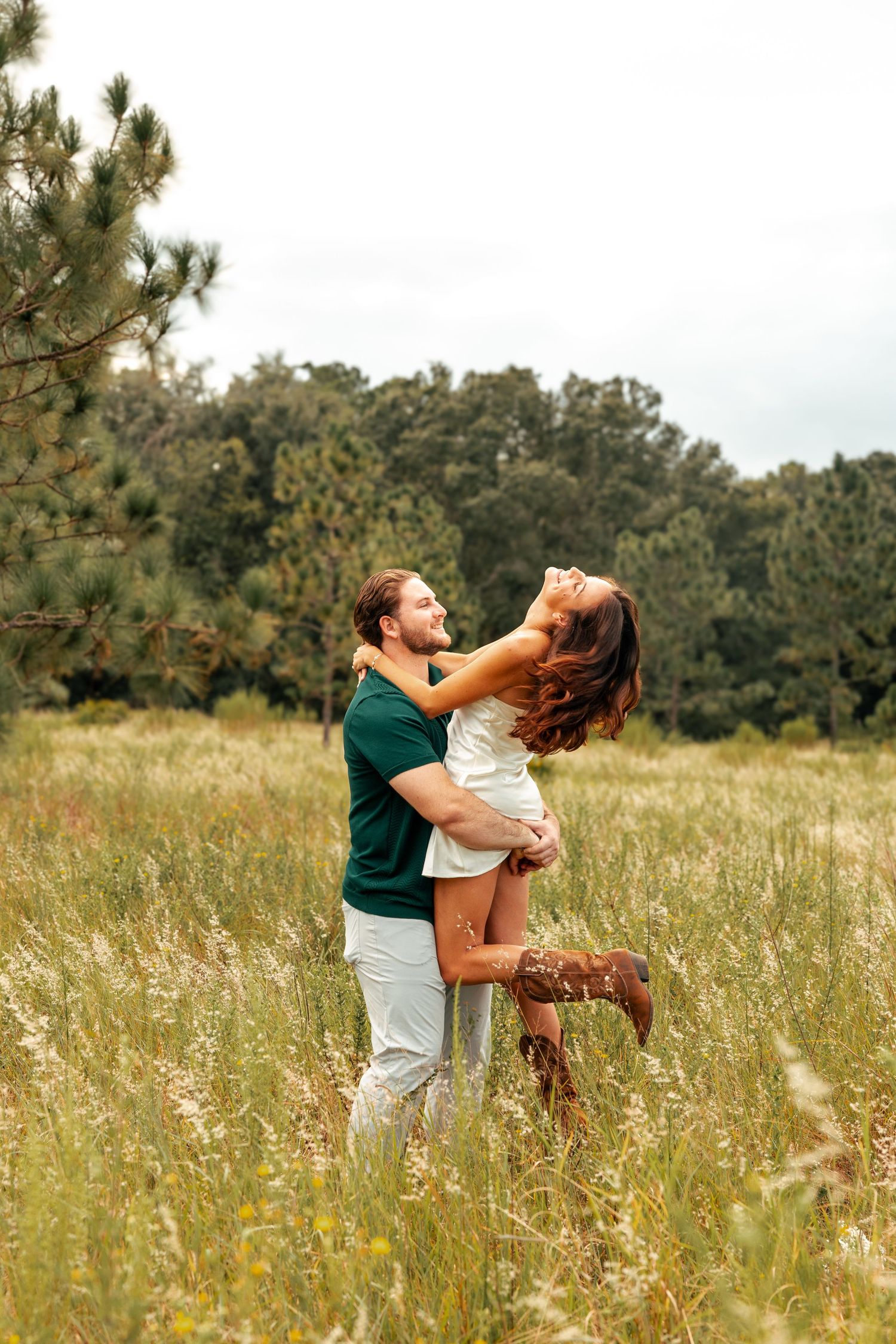 Couple dancing and embracing joyfully in a sunlit meadow filled with tall wildflowers and pine trees in the background.