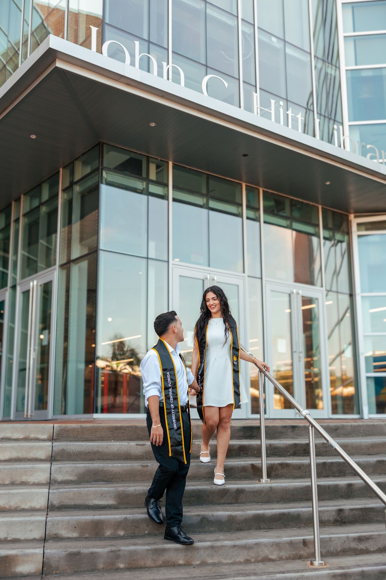 Two graduates in formal attire stand on steps outside modern glass building entrance during graduation photoshoot.