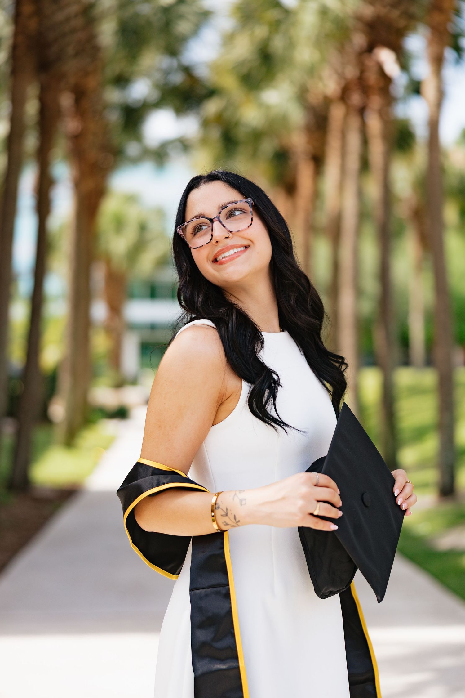 Smiling woman in white dress holding graduation gown outdoors among palm trees, wearing glasses and gold bracelet.