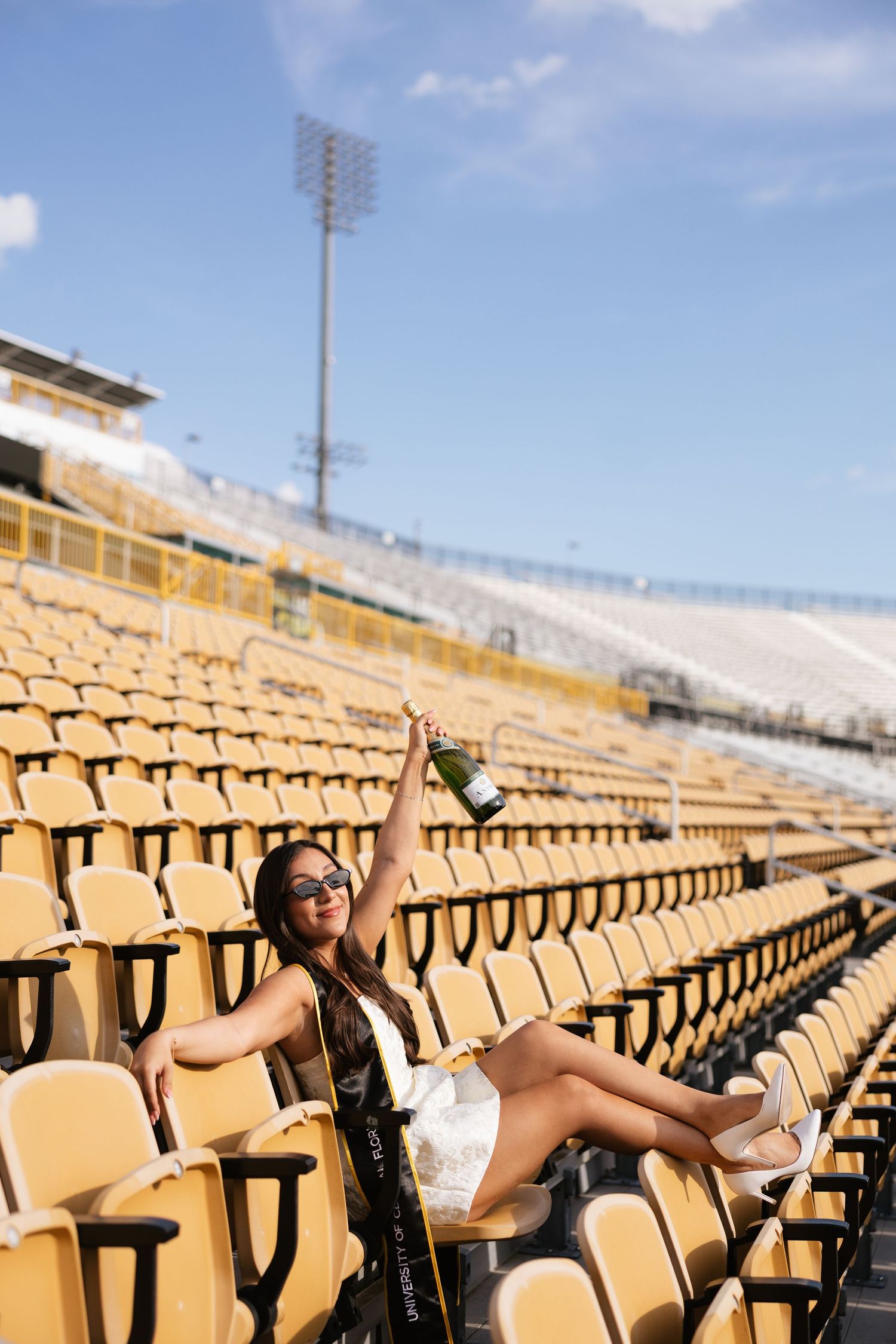Woman in white dress celebrating with champagne bottle in empty yellow stadium seats on a sunny day.