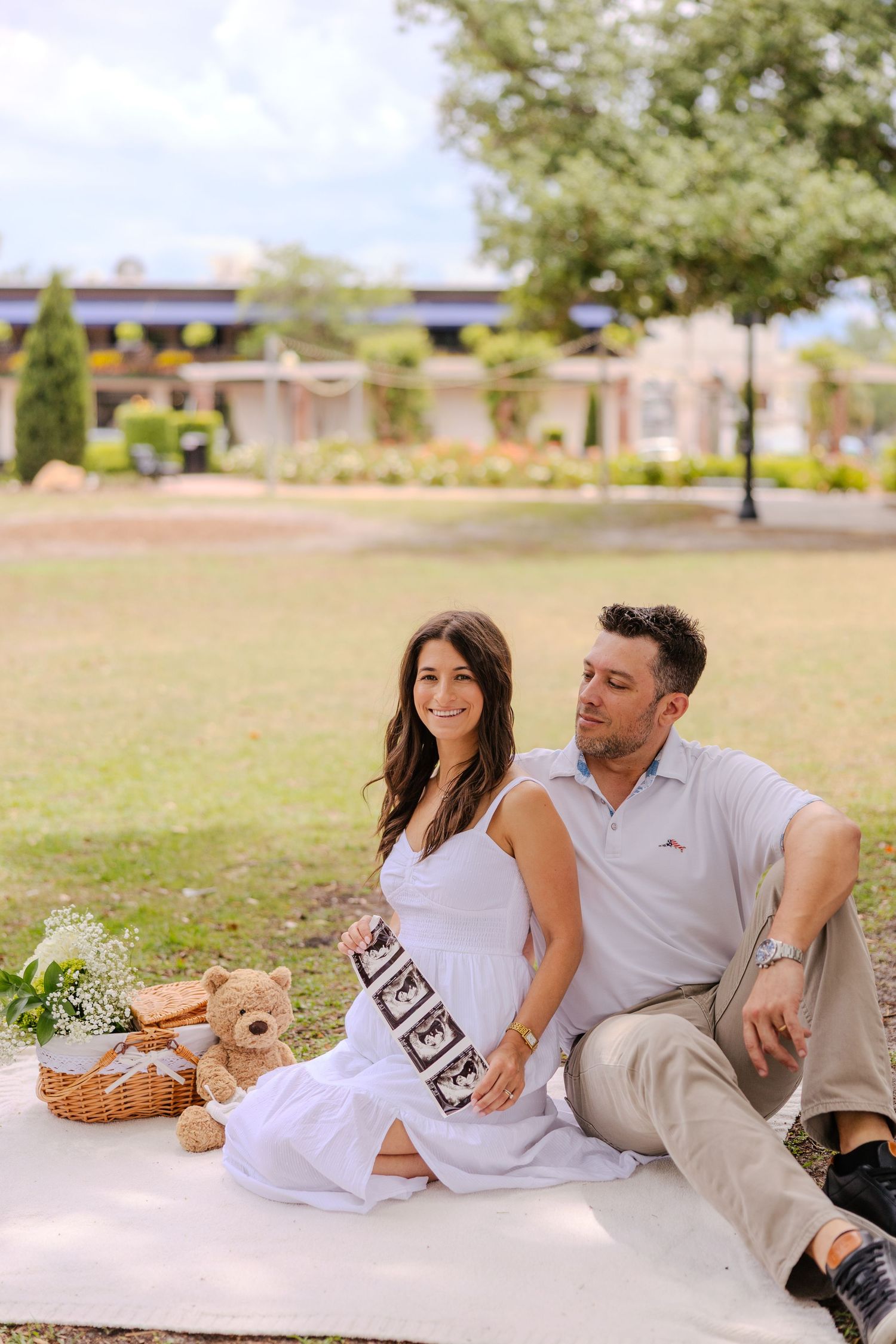 Pregnant couple sitting on blanket in park with ultrasound photos, teddy bear, and picnic basket during maternity photoshoot.