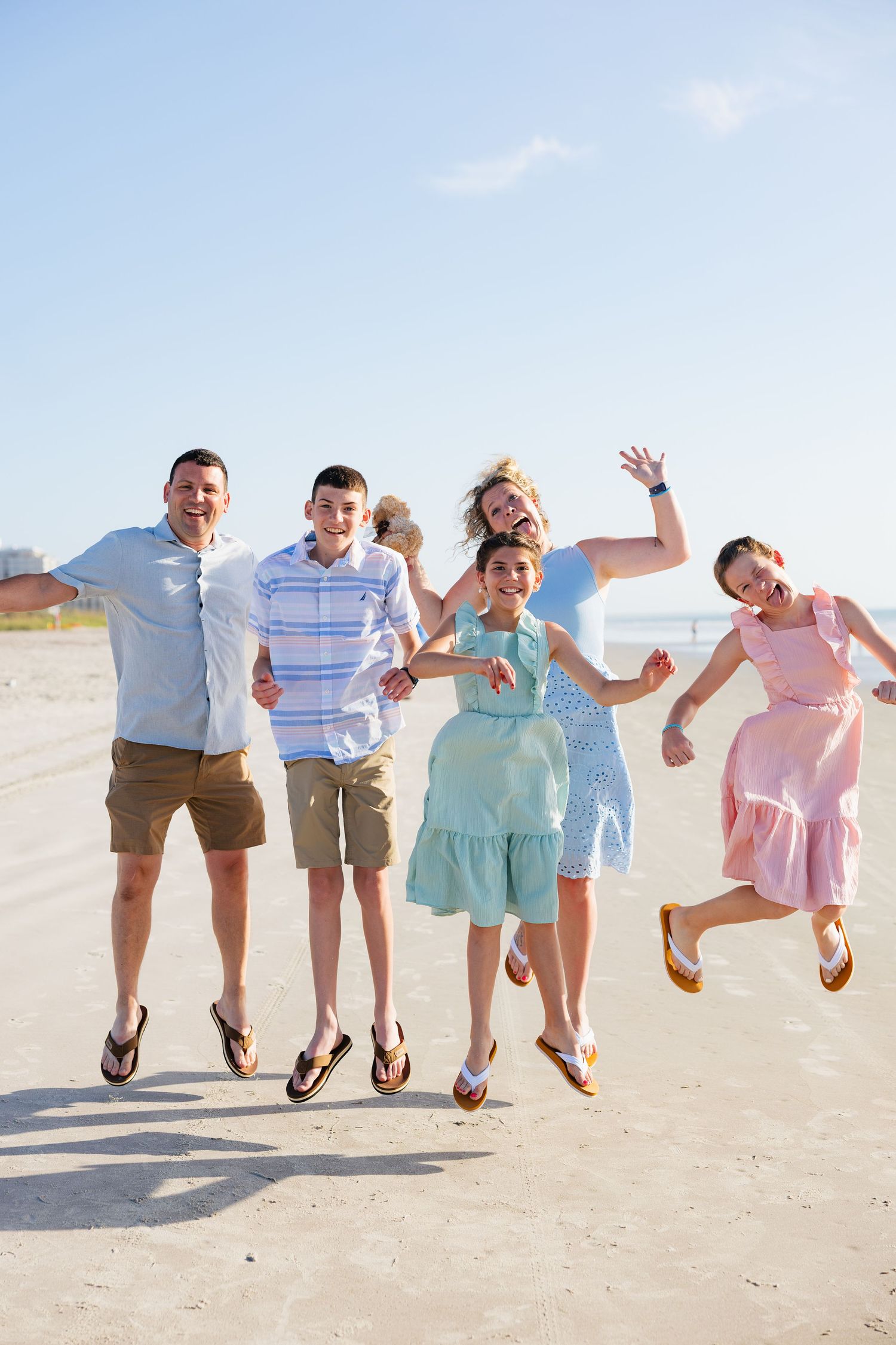 Happy family of six jumping together on a sunny beach, wearing casual summer outfits, smiling and waving at the camera.