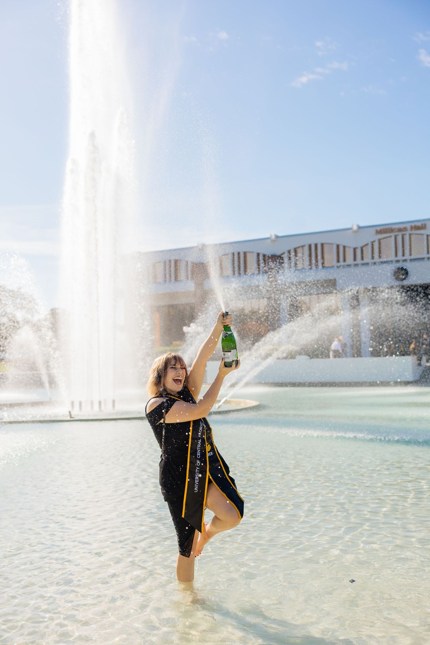 A person in a black dress celebrates with a champagne bottle in a fountain on a sunny day.
