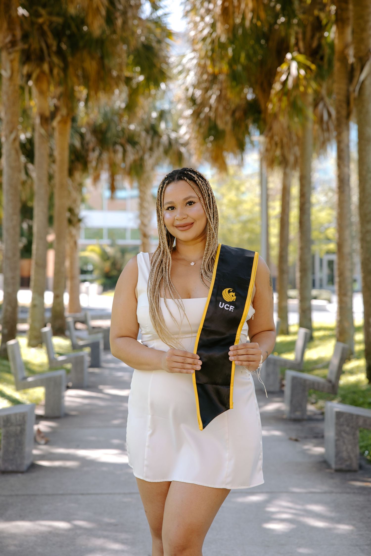 Graduate in white dress poses with graduation sash on tree-lined pathway.