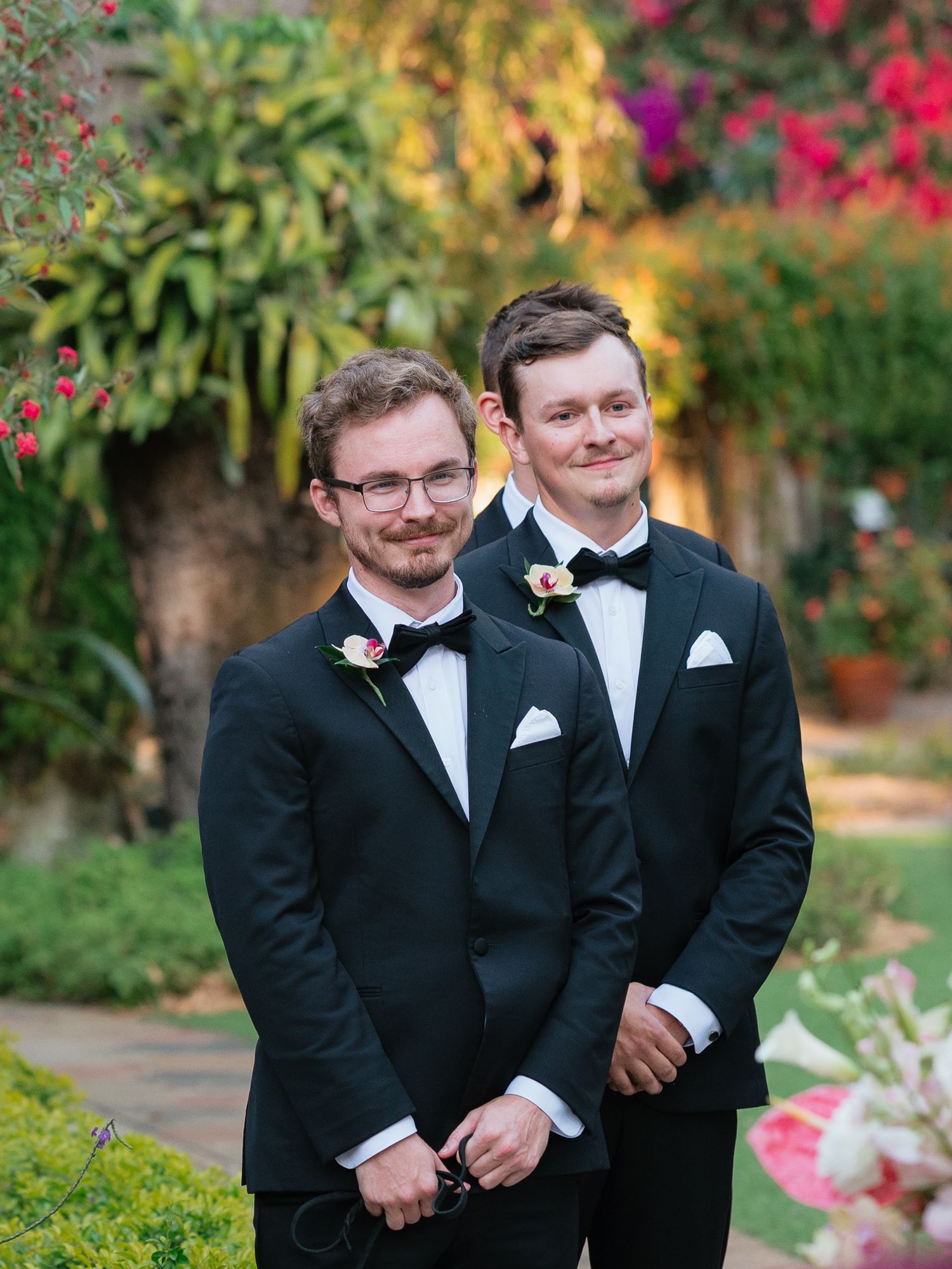 Two people in black tuxedos and bowties share a candid moment in a garden with colorful flowers in the background.