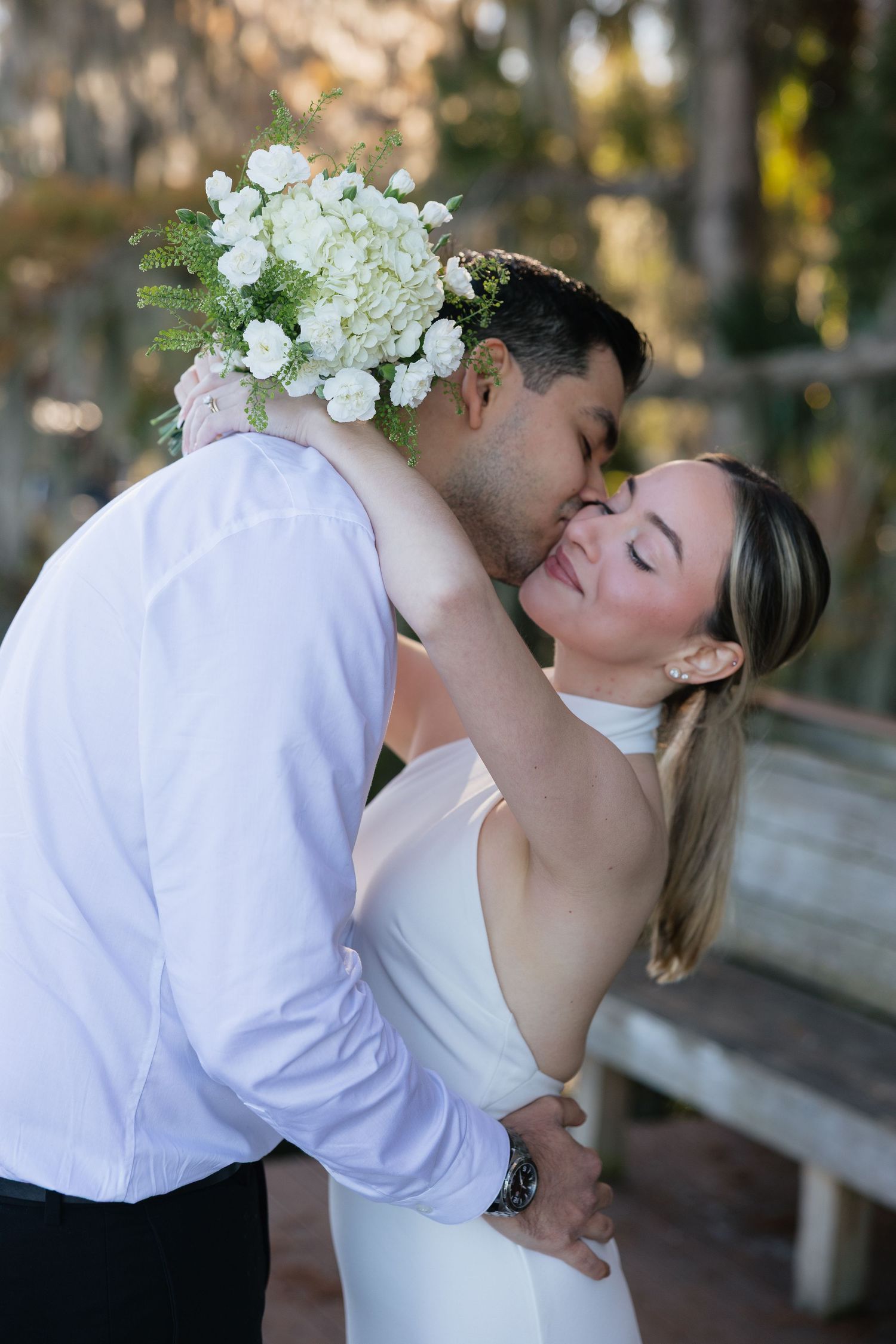 A romantic wedding couple shares tender kisses in a garden setting wearing white attire.