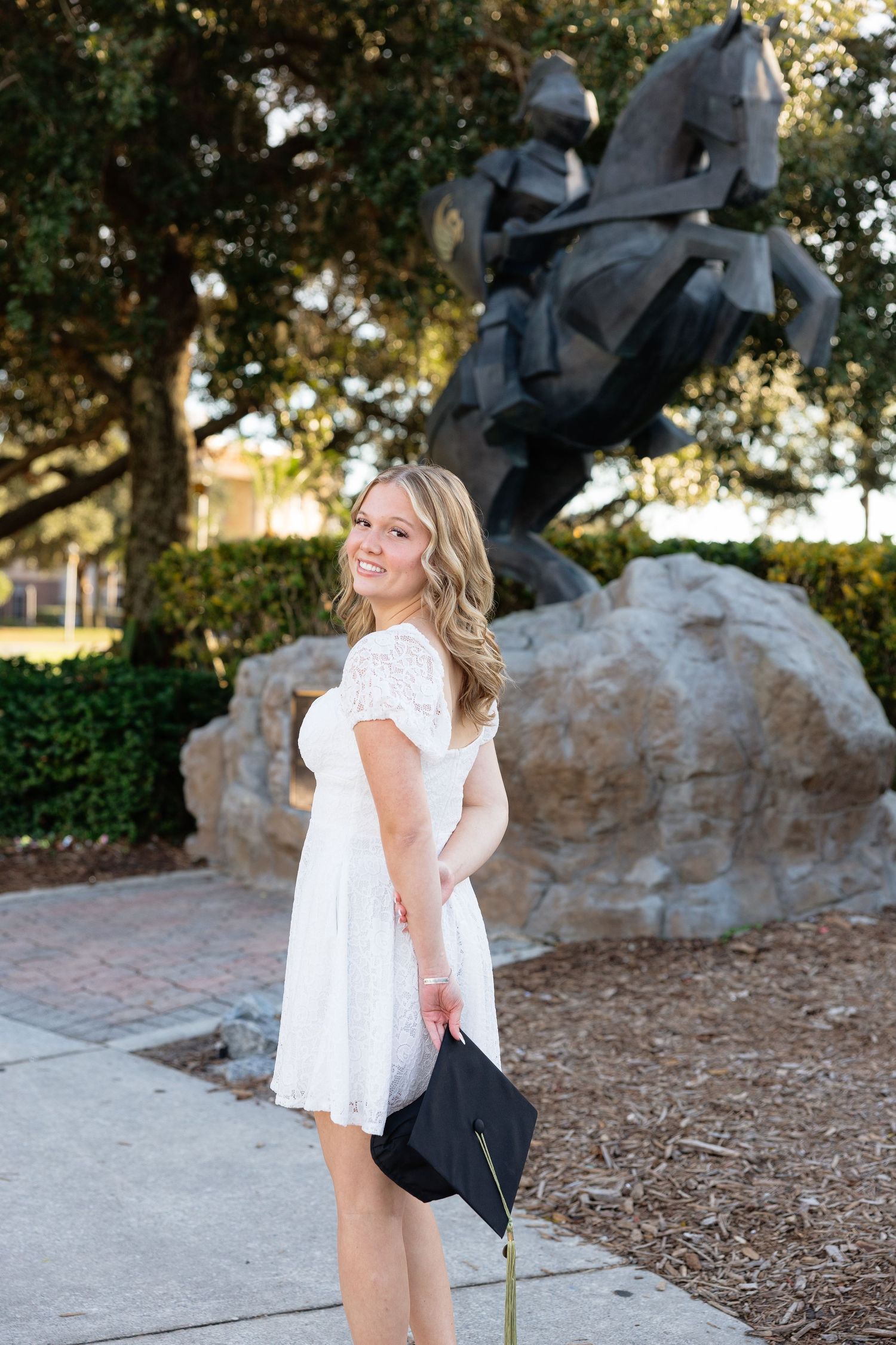 A figure in a white dress poses near a stone sculpture in a garden setting during golden hour lighting.