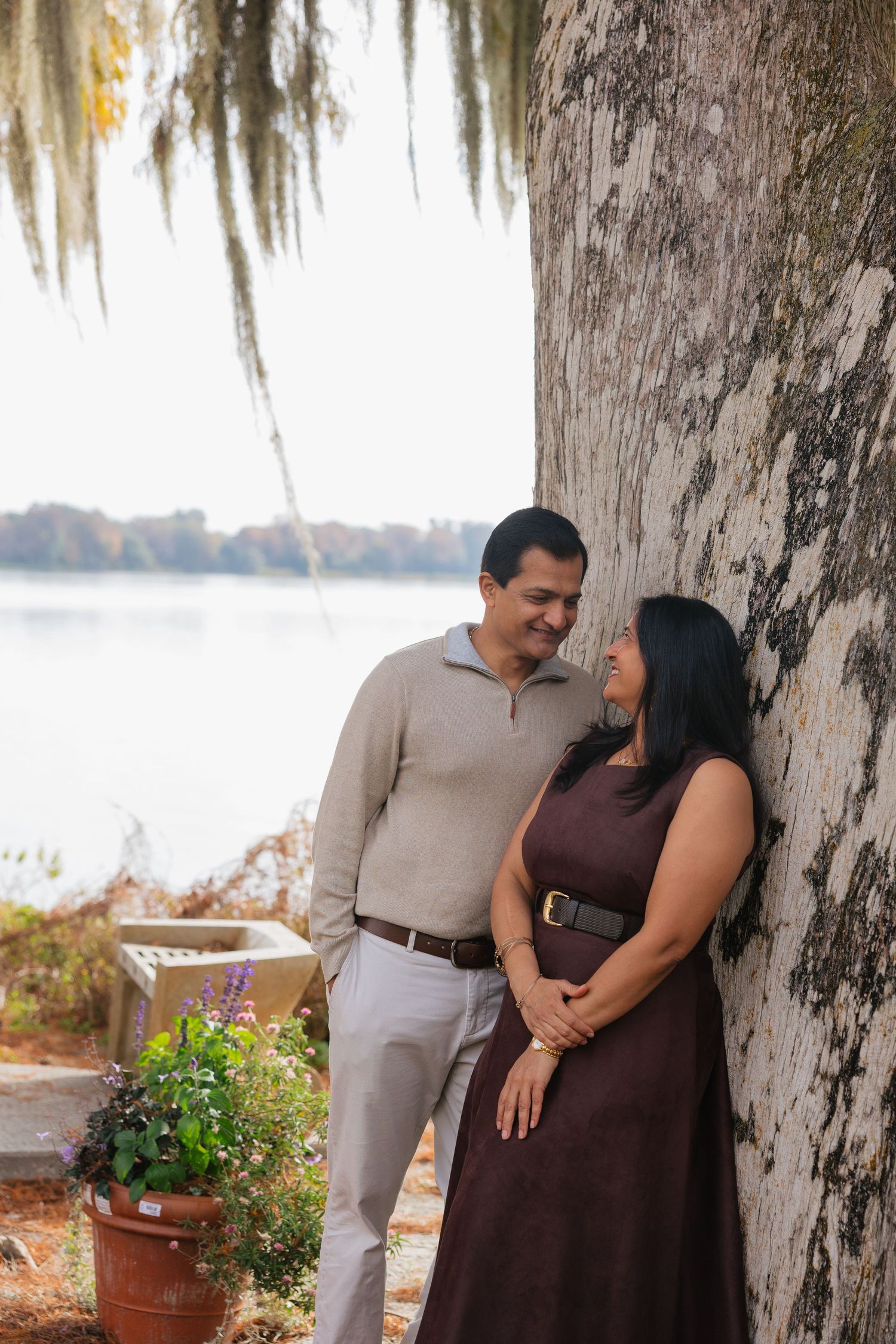 A couple shares an intimate moment beside a large tree trunk near a lake with Spanish moss hanging overhead.