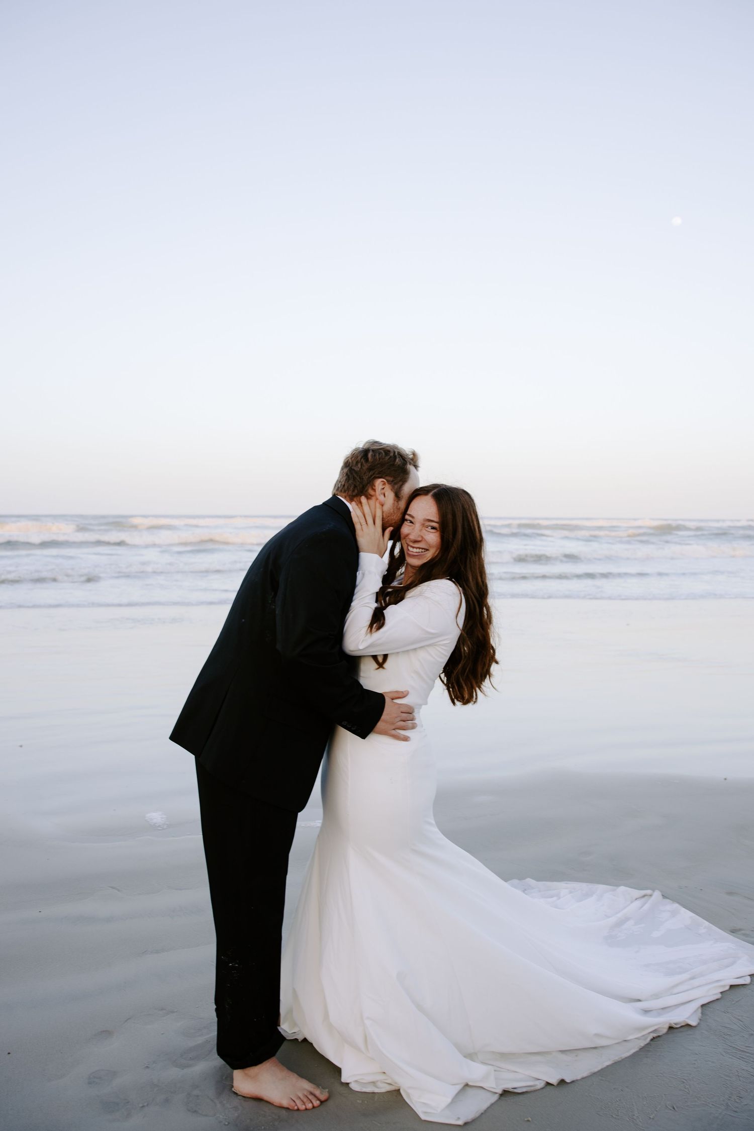 Beach wedding photoshoot with couple dancing and embracing on sandy shore at sunset.