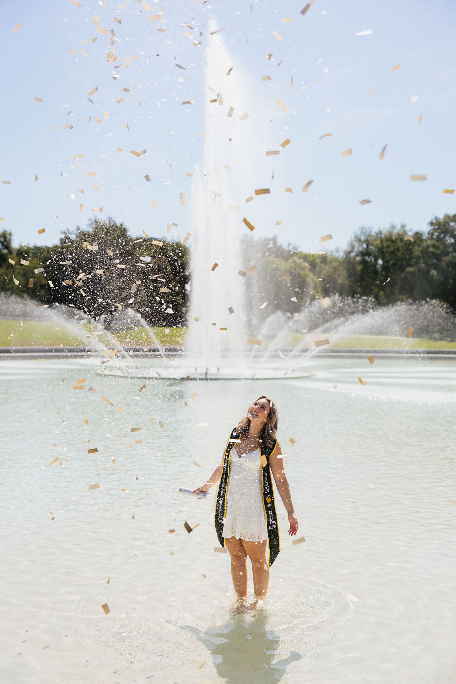 Graduate in white dress and sash celebrates with gold confetti at a fountain on a sunny day.