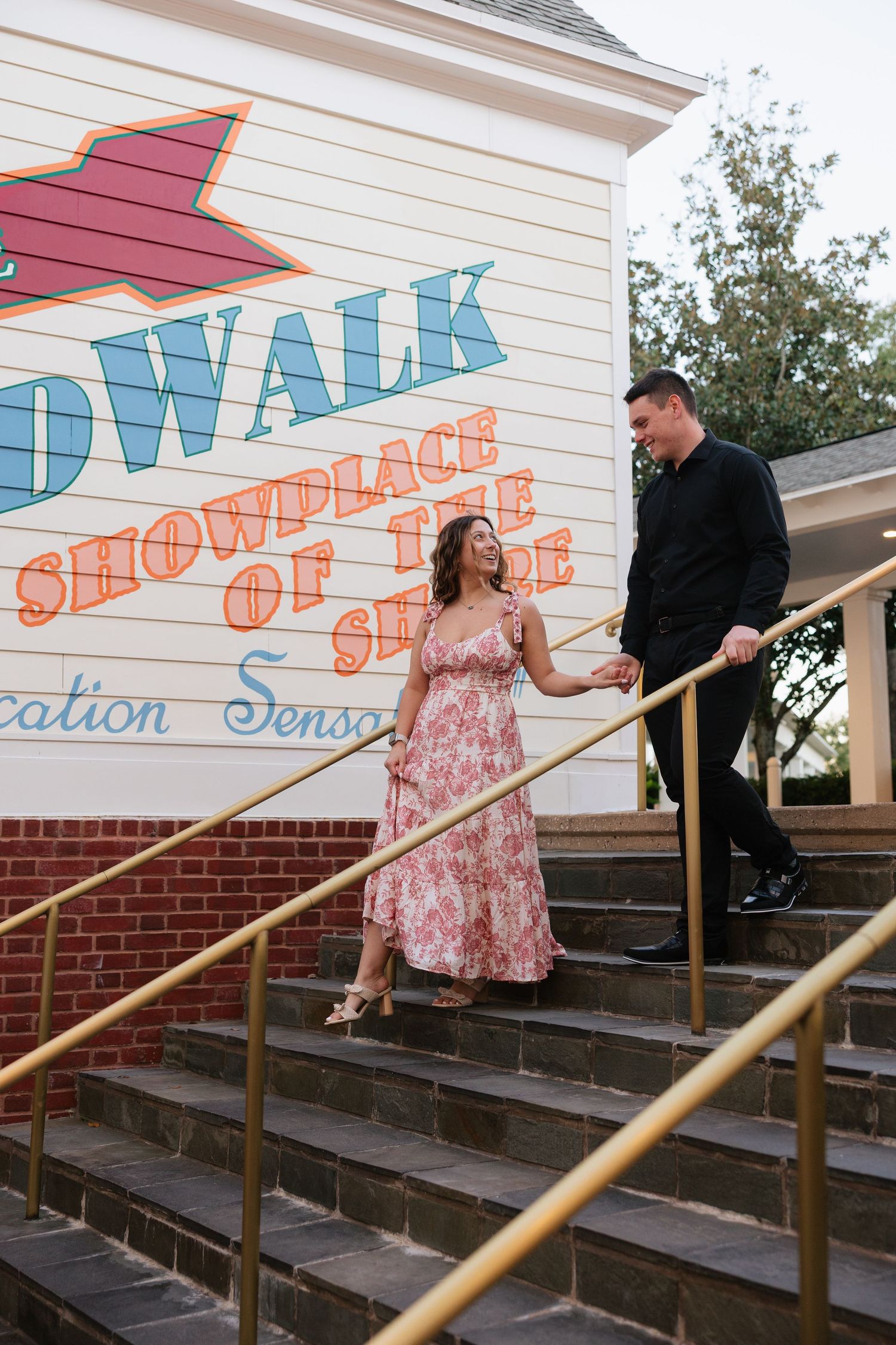 A couple shares a lighthearted moment on brick steps with a handrail outside a building with vintage signage.