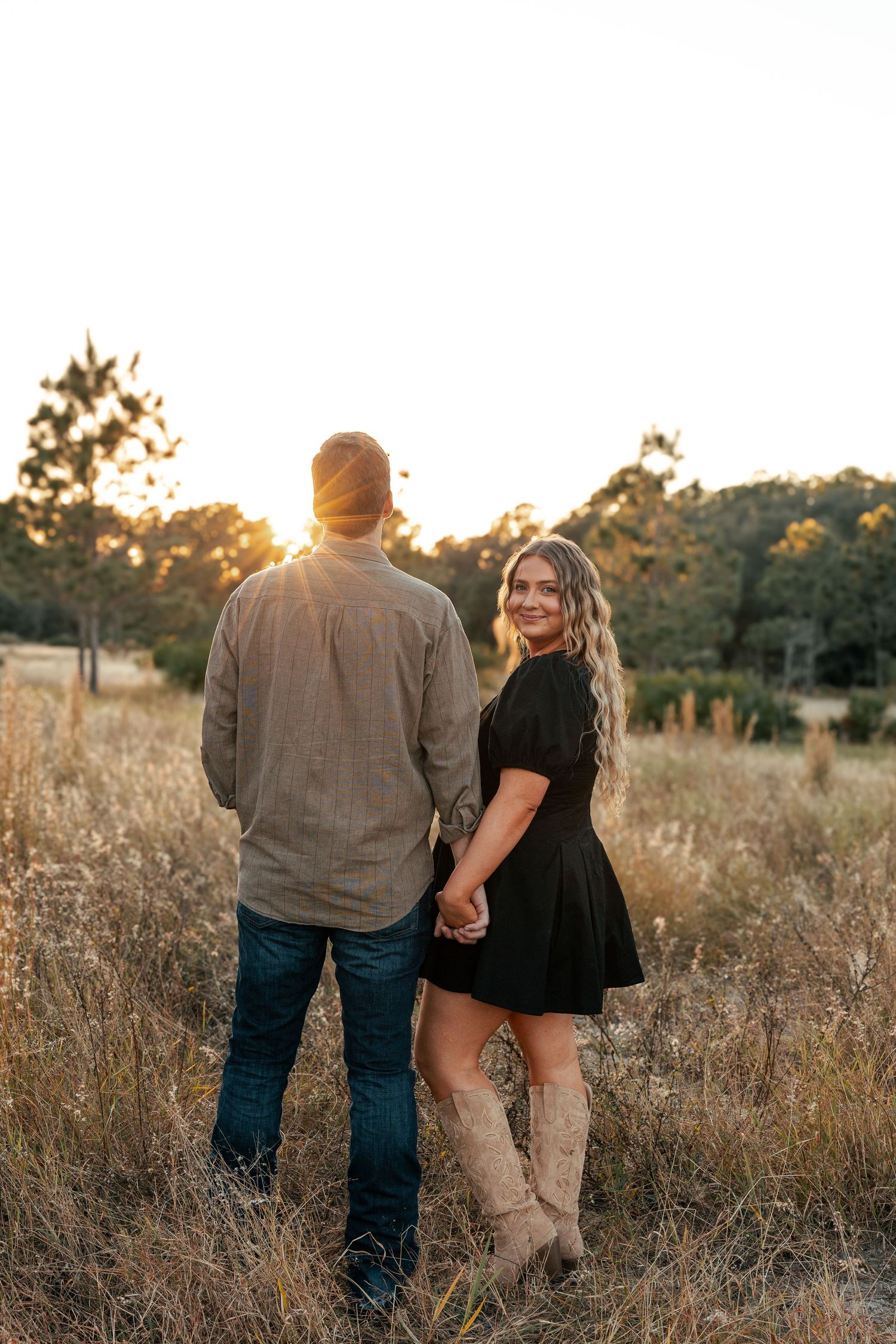 A couple holds hands in a golden field at sunset while wearing casual attire and cowboy boots.