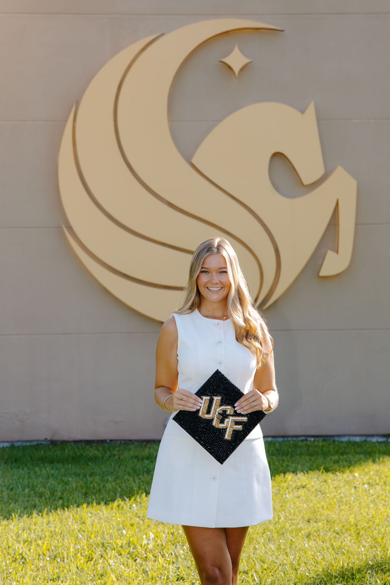 Graduating student poses in front of university logo wearing white dress and holding graduation cap.