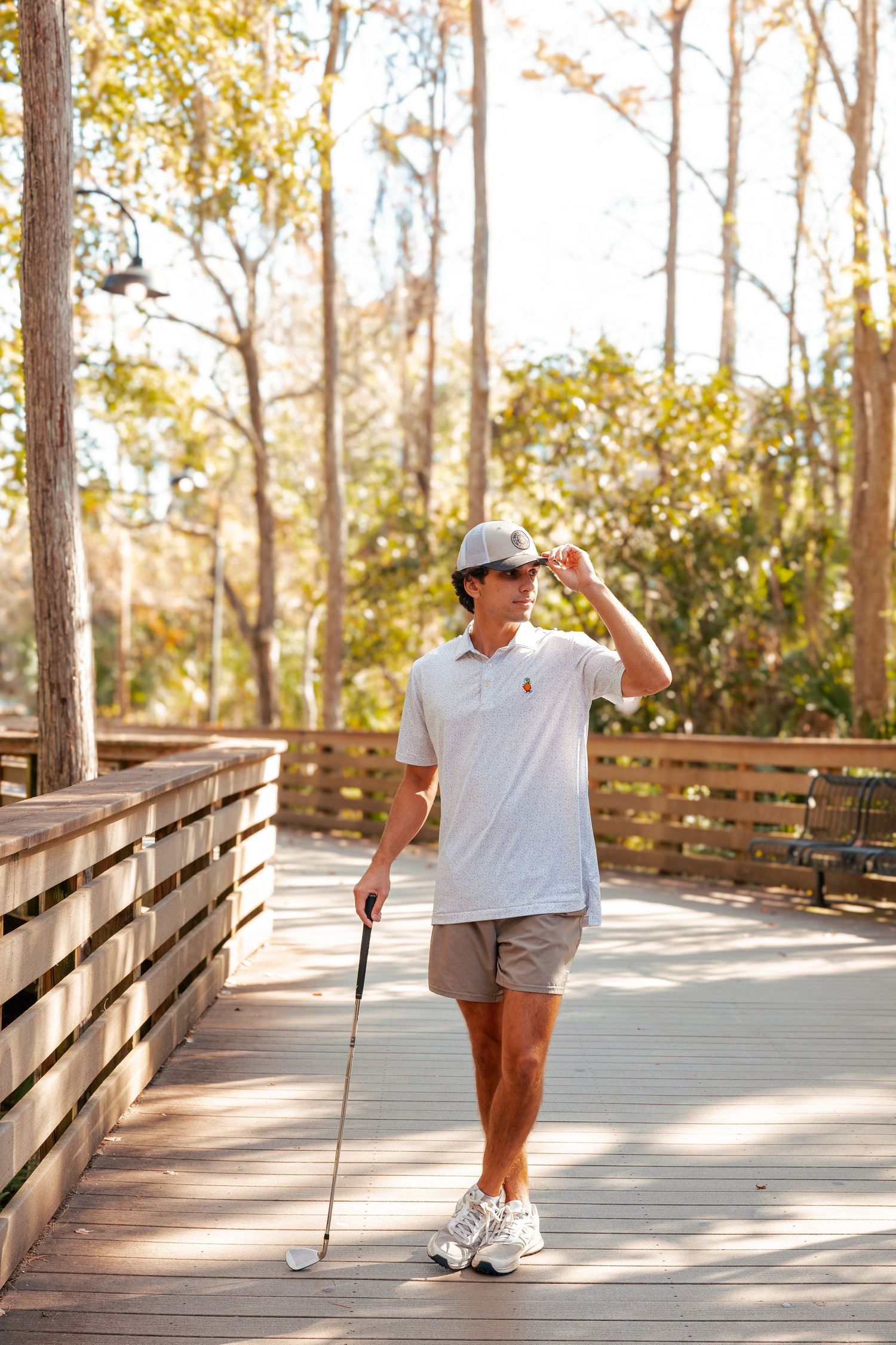 Golfer in white polo and shorts stands on wooden boardwalk path holding golf club on sunny day.